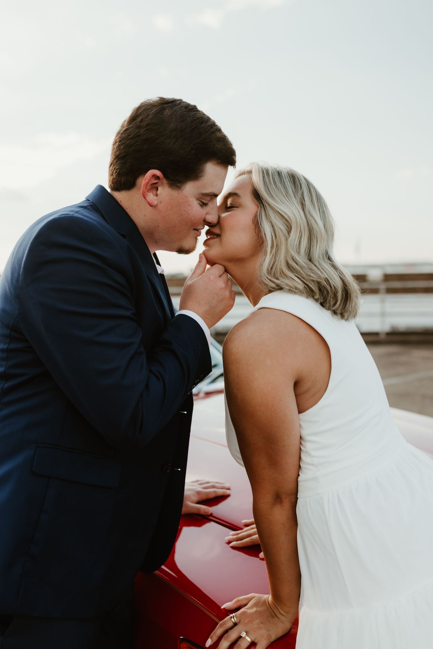 A romantic moment between a couple in wedding attire leaning against a red car at sunset.
