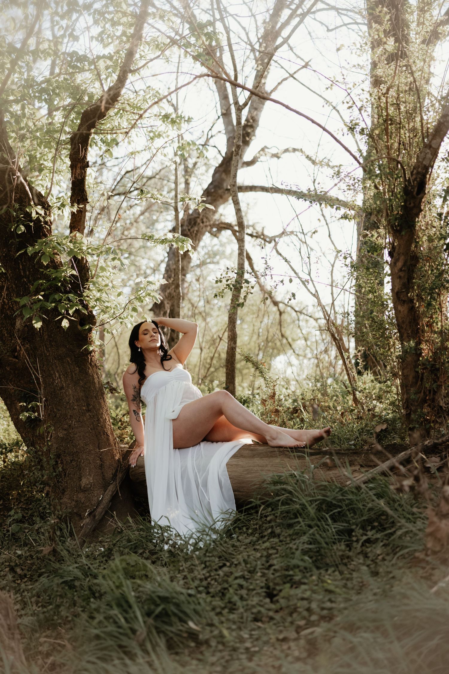 Ethereal maternity portrait in a white dress against sunlit forest trees.