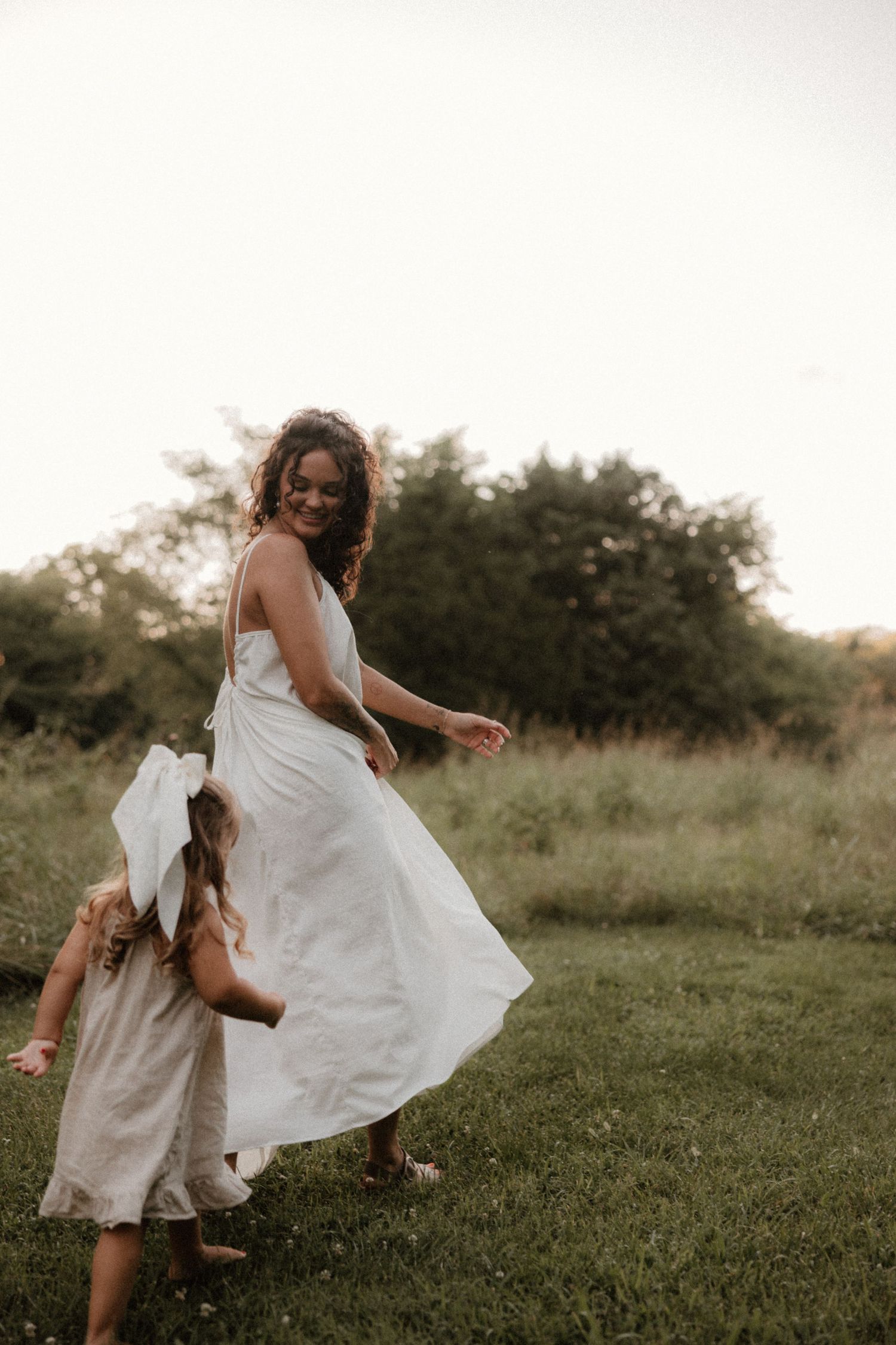 Two people in white dresses dance joyfully together in a grassy outdoor field at sunset.