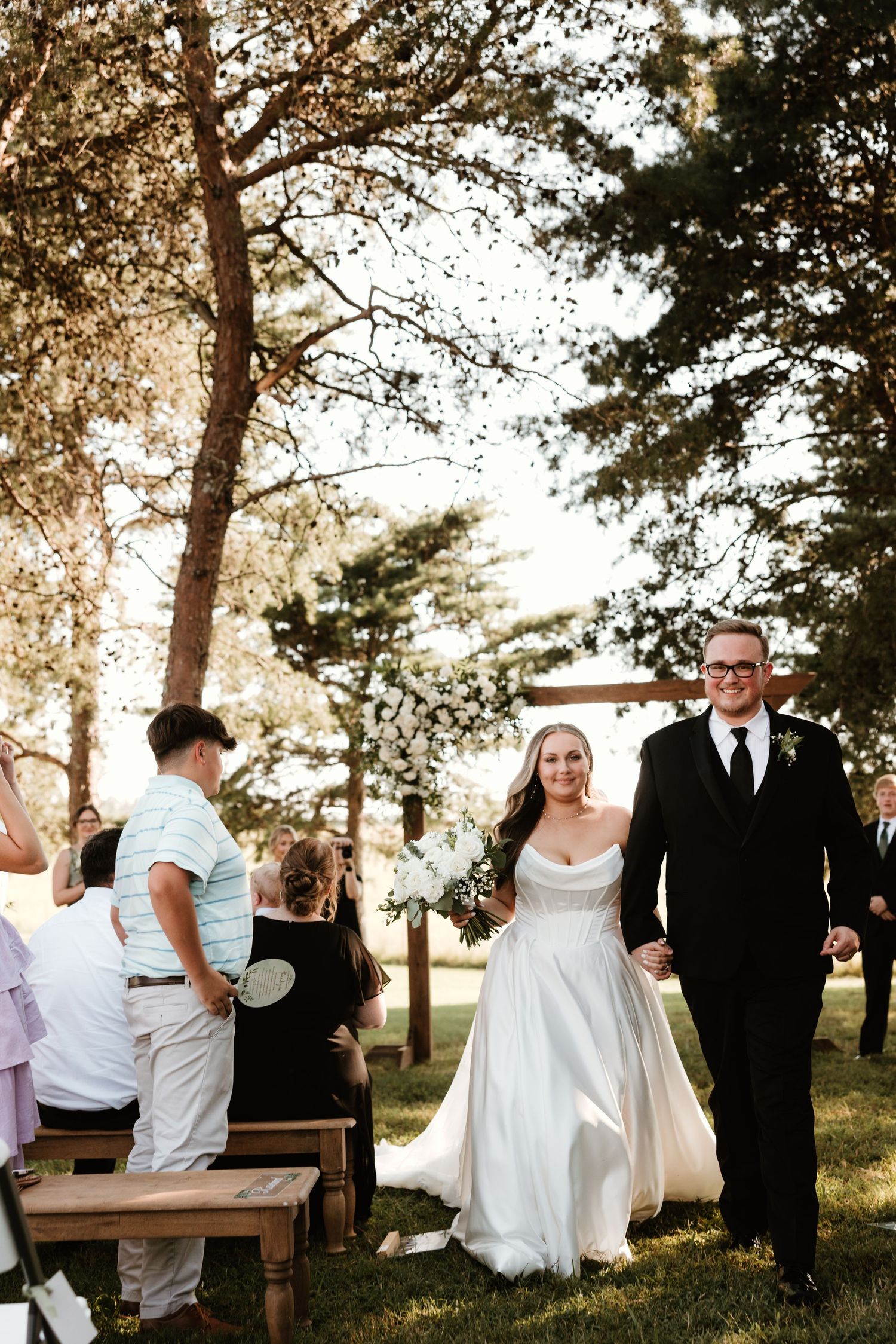 A romantic outdoor wedding ceremony in a wooded setting with sunlight filtering through tall pine trees.