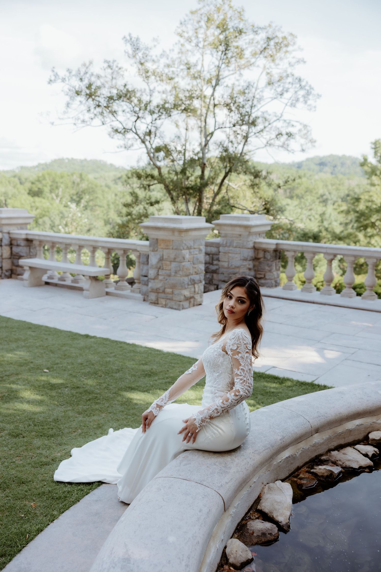 Bride in lace long-sleeve wedding gown seated on stone fountain edge at elegant outdoor venue with lush greenery.