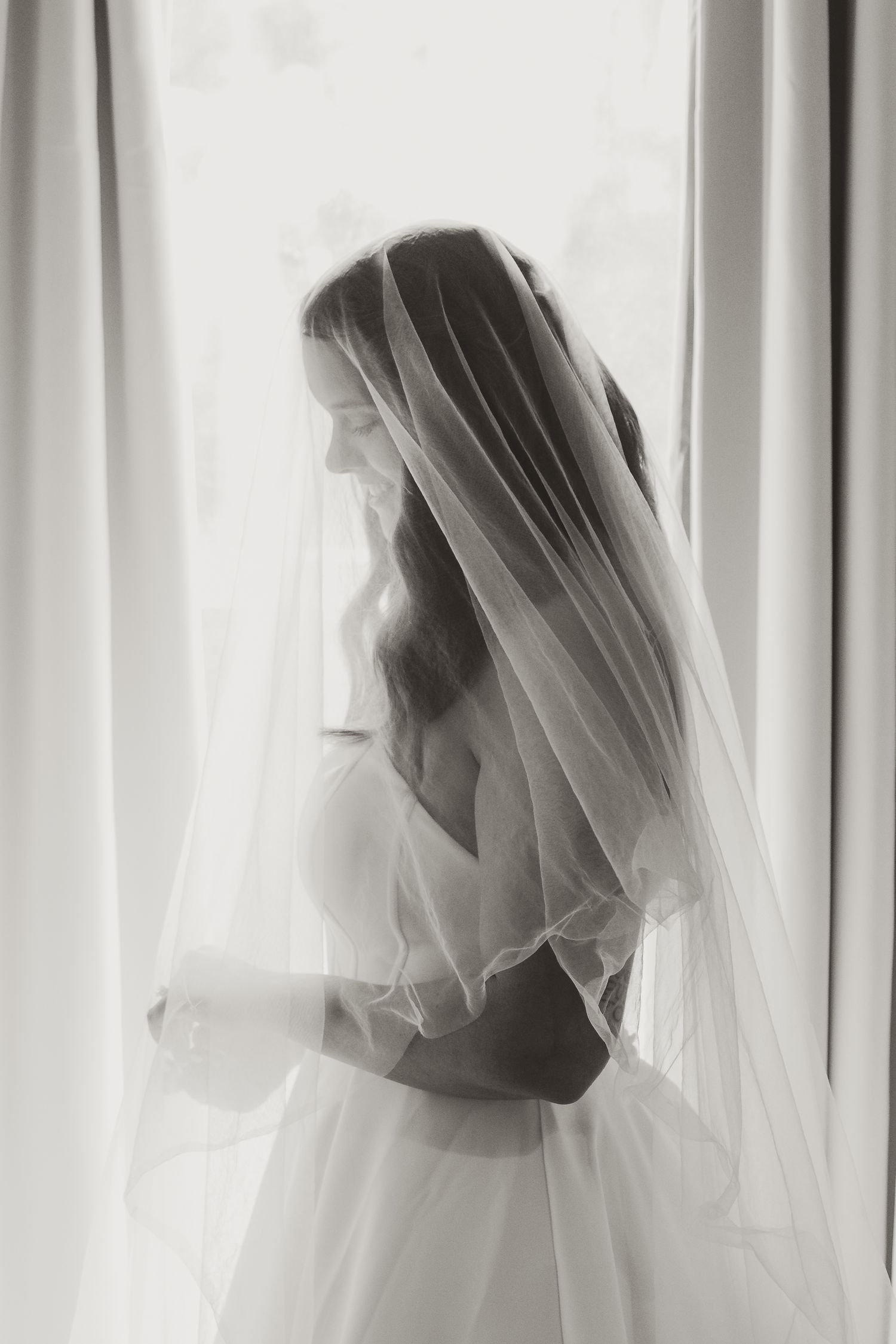 Black and white photo of a bride in a wedding dress and veil standing by a window in soft natural light.