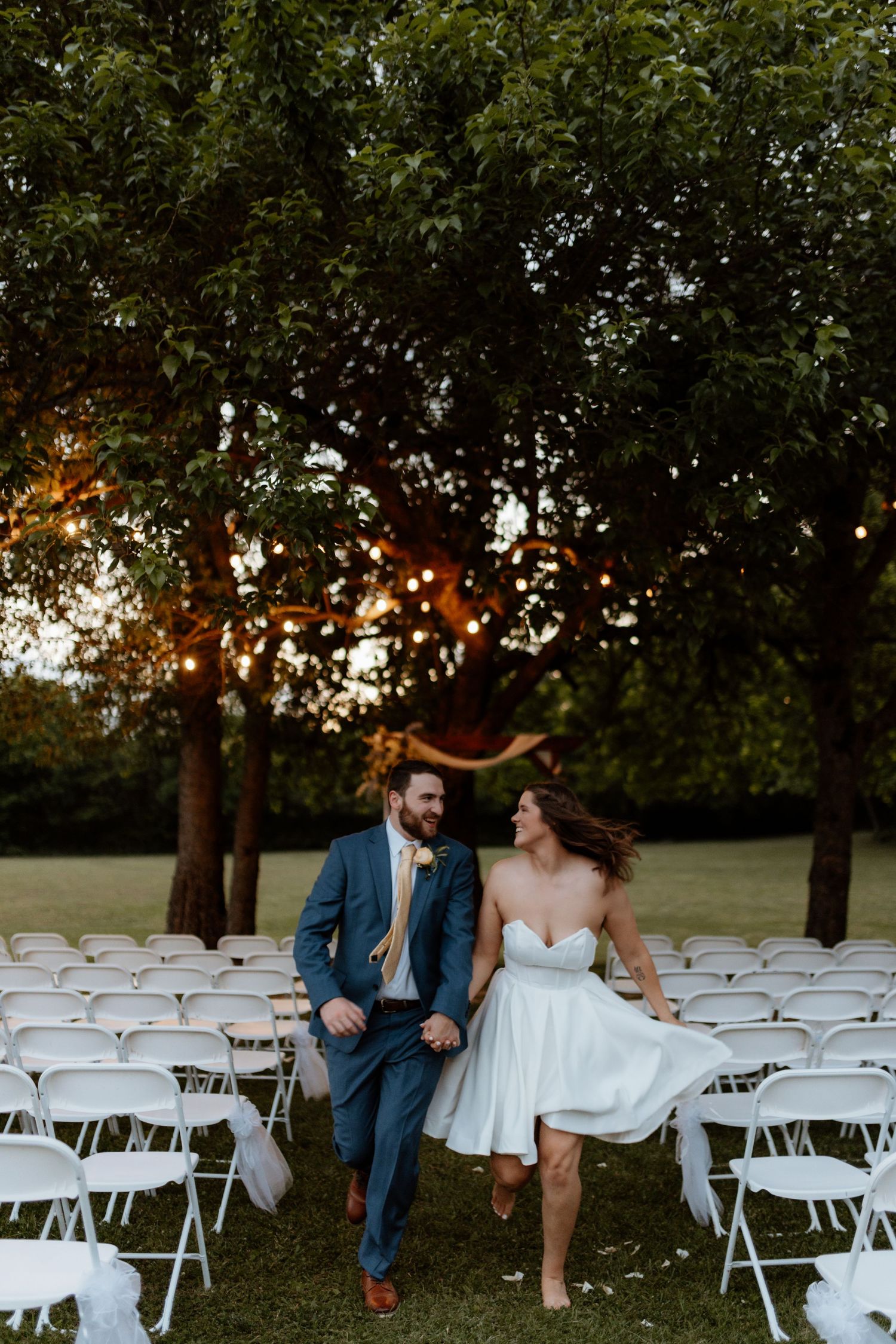 Bride and groom walk hand-in-hand through outdoor wedding venue with white chairs and string lights at sunset.