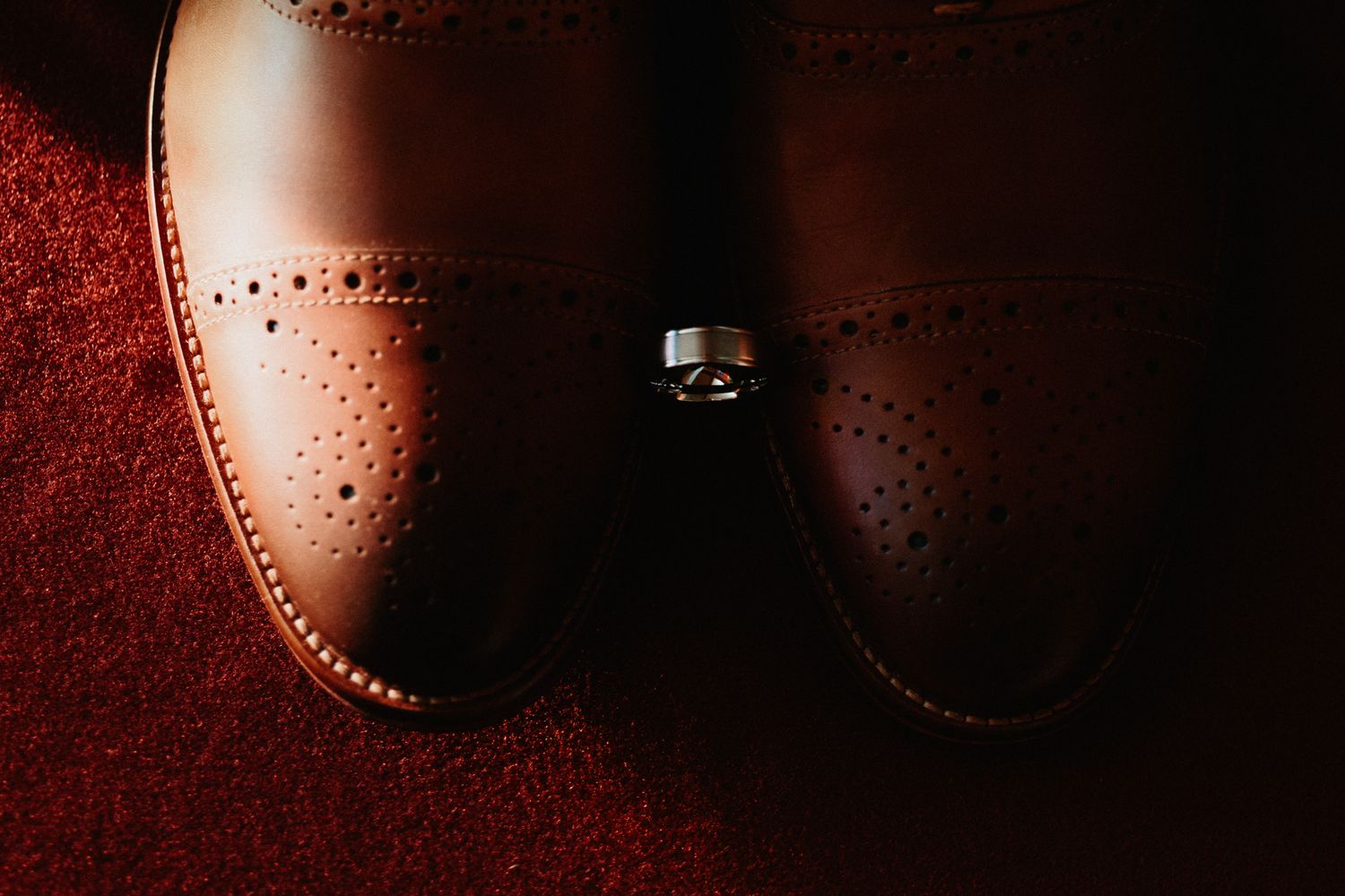 A silver wedding ring sits atop brown leather dress shoes against a dark red background.