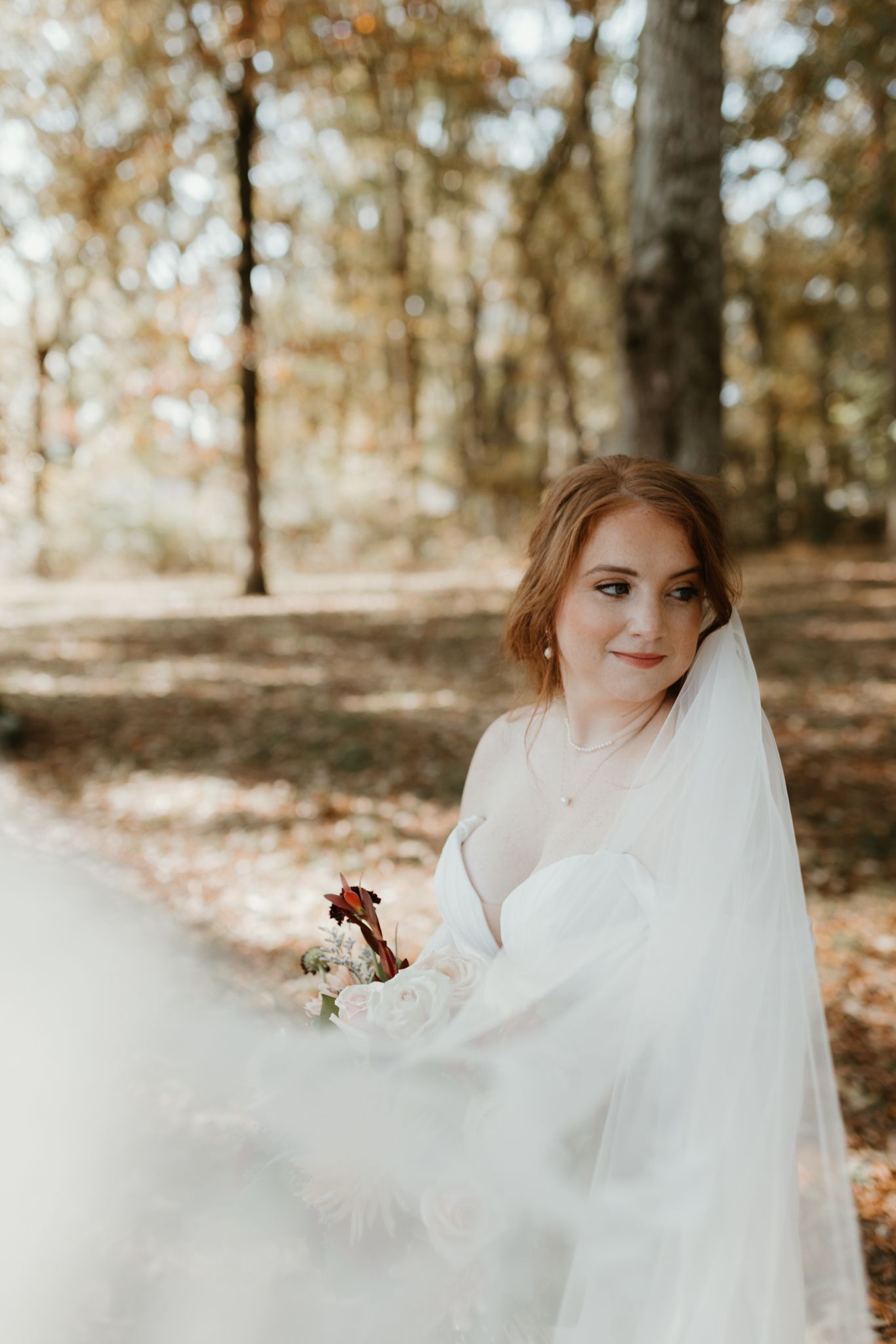 A person in a flowing white wedding dress twirls gracefully among autumn trees with fallen leaves on the ground.