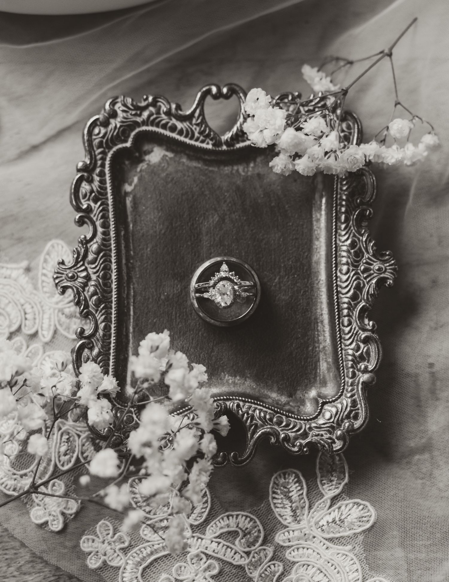 An ornate silver frame adorned with delicate lace and floral elements surrounds a vintage ring in black and white.