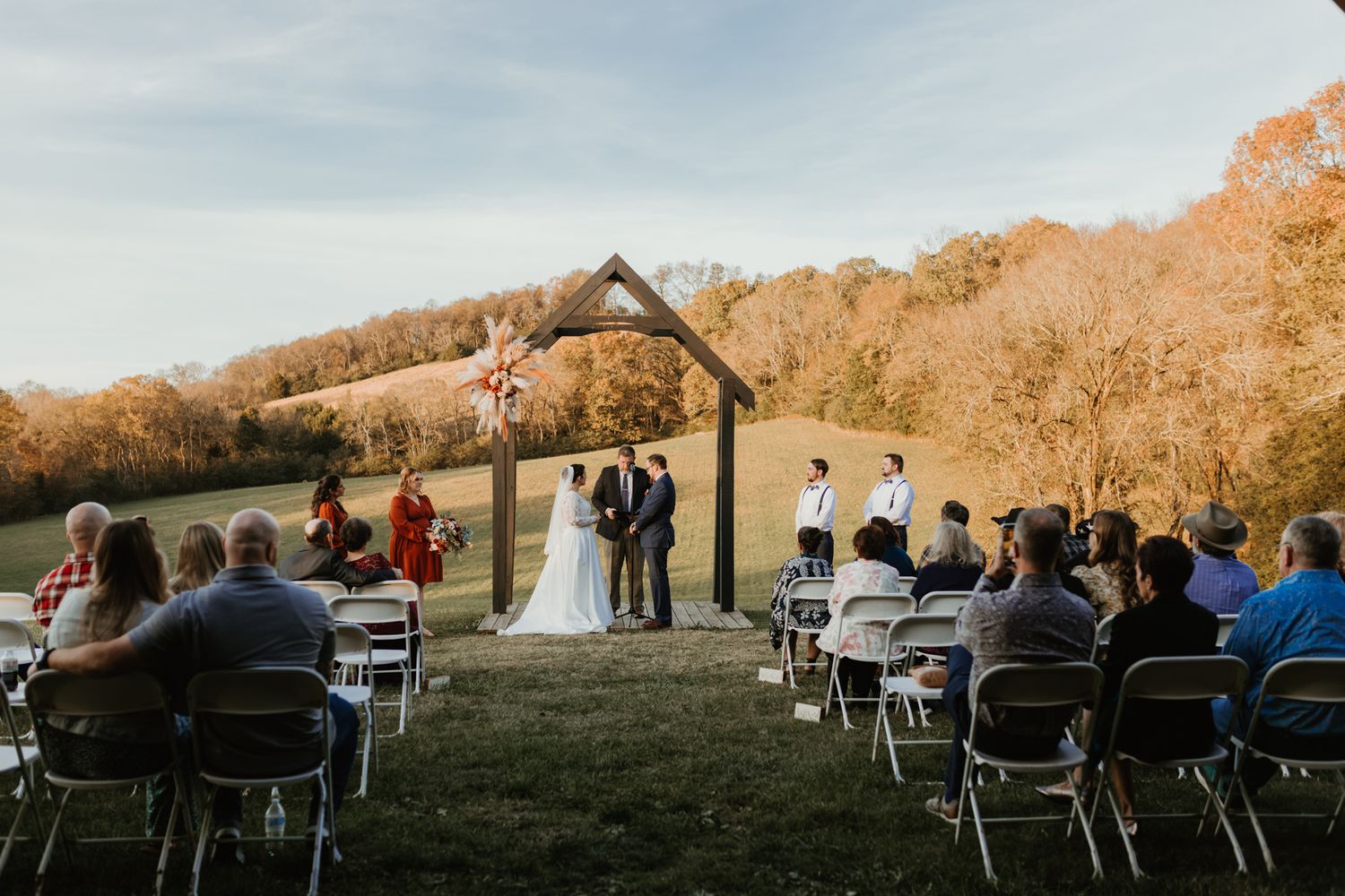 Guests seated in white chairs on a grassy hillside for an outdoor wedding ceremony at sunset.
