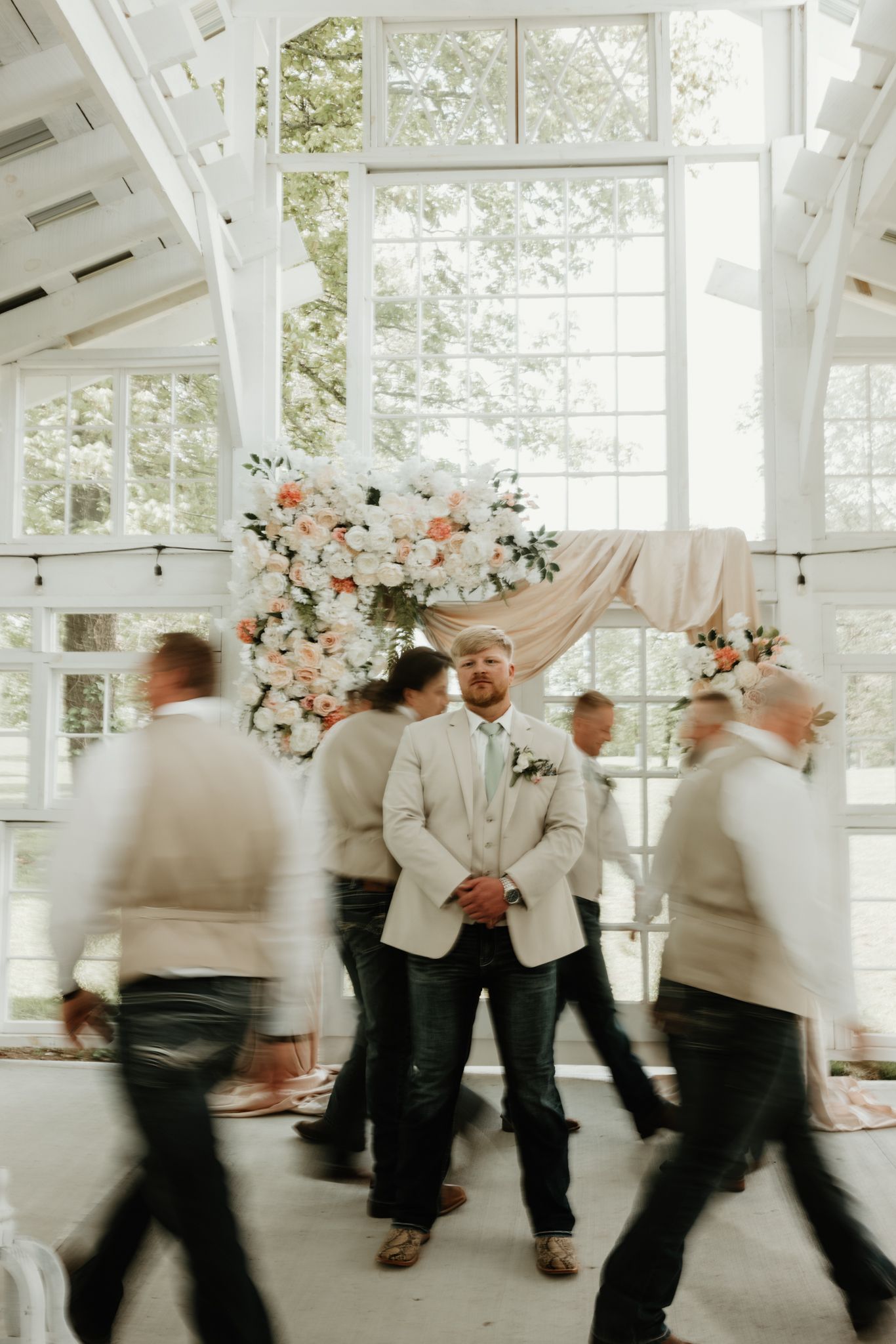 Motion-blurred wedding guests dance and move in a sunlit reception venue with floral decorations.
