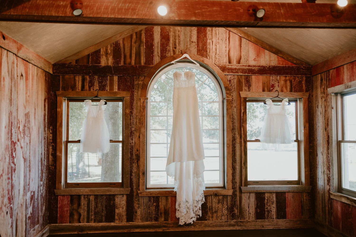 White wedding gown hangs in center window of rustic wooden cabin with sunlight filtering through.