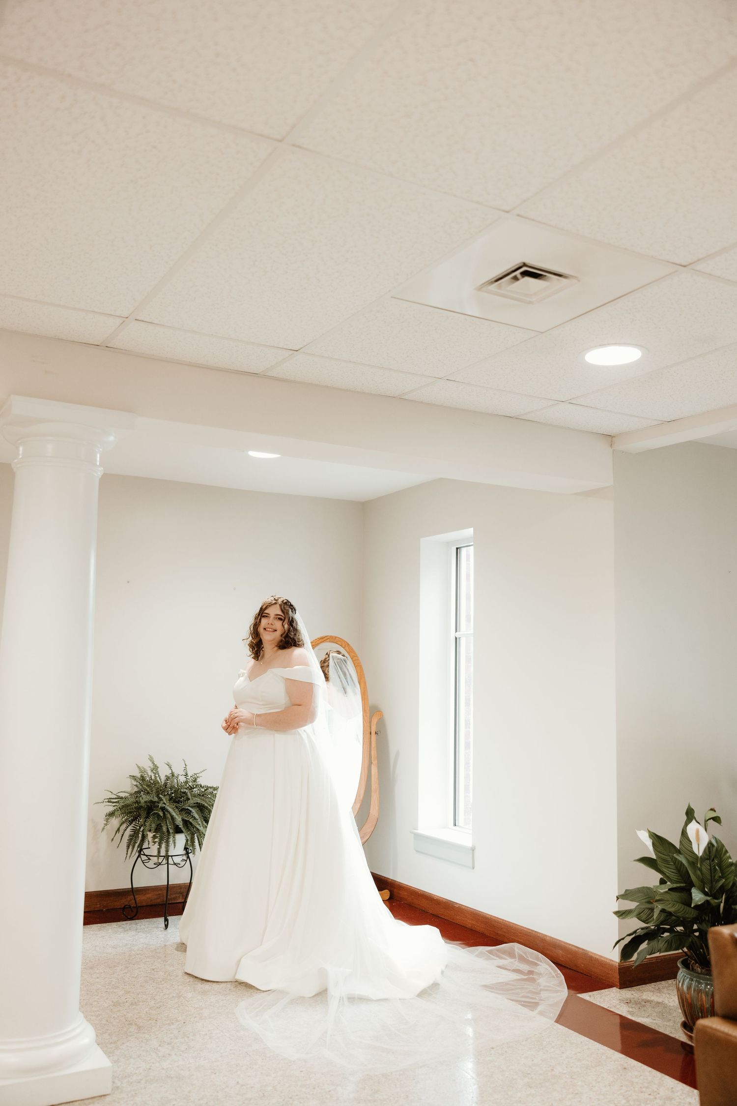 A bride in an elegant off-shoulder white wedding gown stands by a window in a bright interior space with white columns.