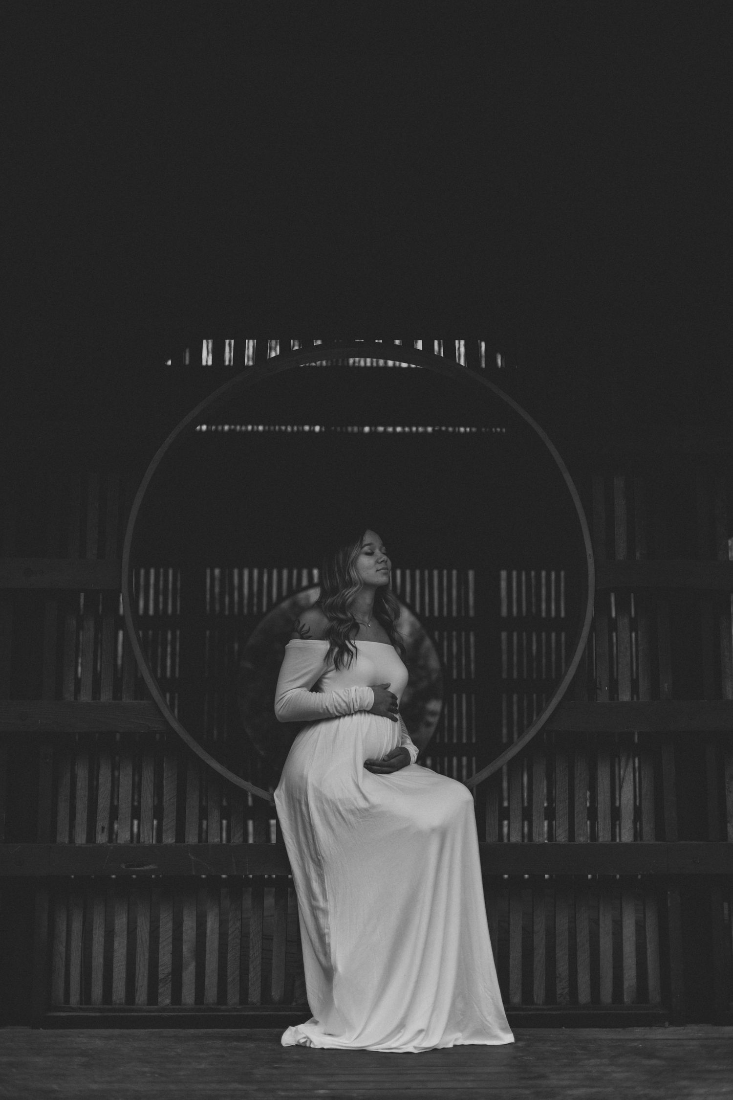 A figure in a white dress sits on a swing in a dimly lit rustic barn setting, creating a moody artistic portrait.