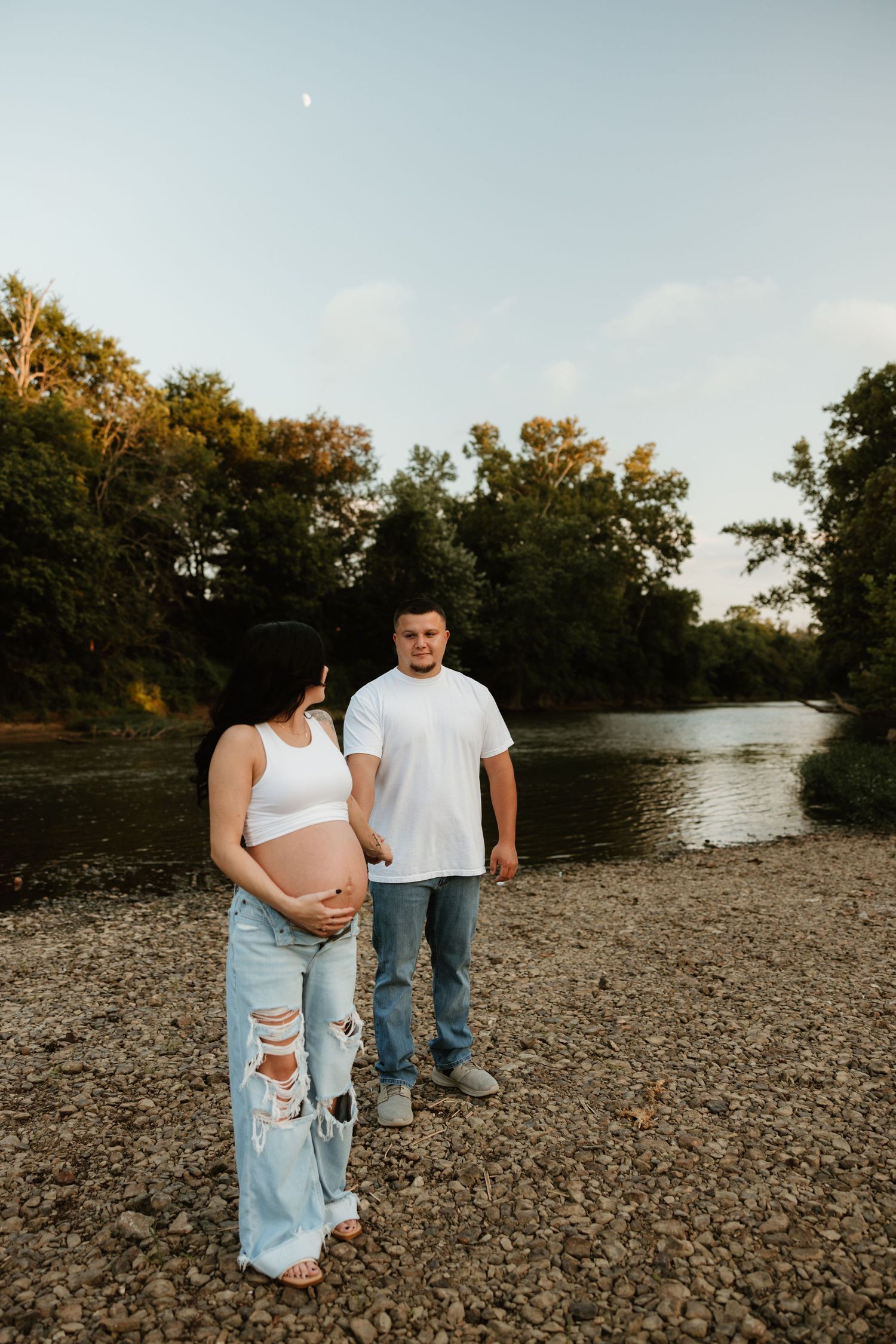 Expecting couple standing on rocky riverbank at sunset with autumn trees in background.