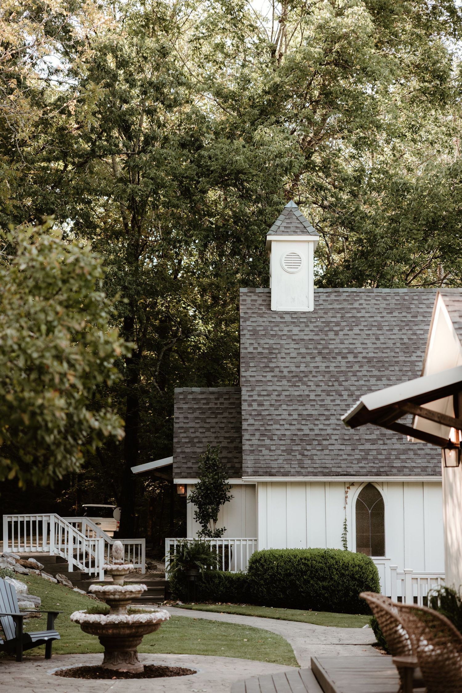 White chapel building with grey shingle roof and cupola nestled among trees in a serene outdoor setting.
