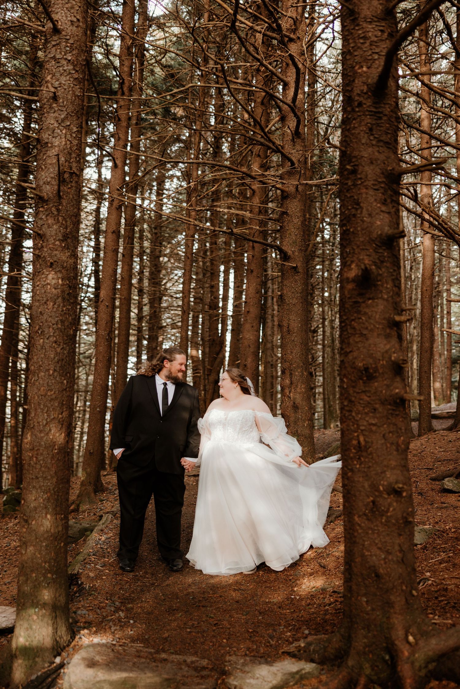 Bride and groom walk hand in hand through a forest, her white gown flowing, as they share a loving glance.