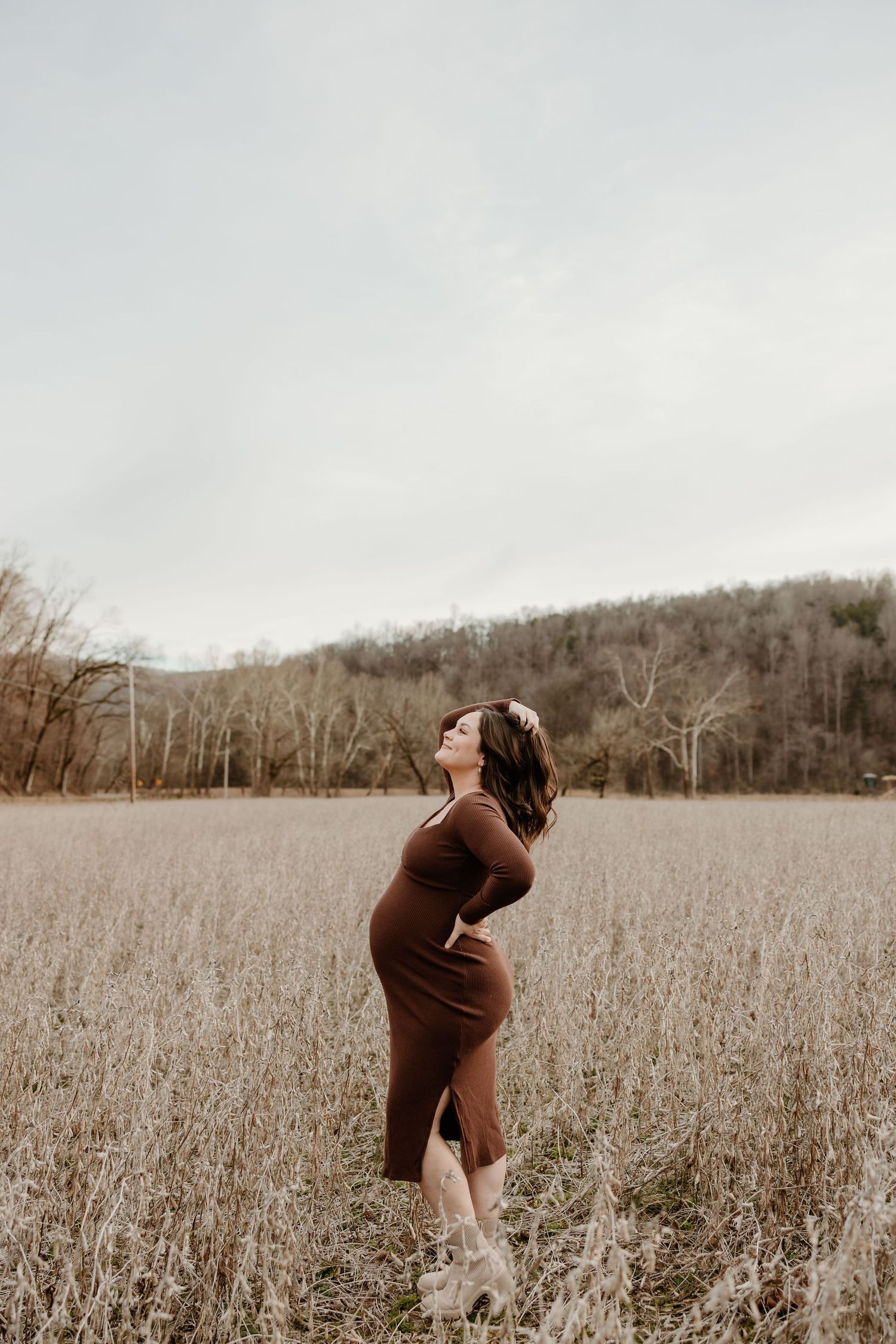 Pregnant woman in brown dress standing in autumn field with trees and overcast sky.