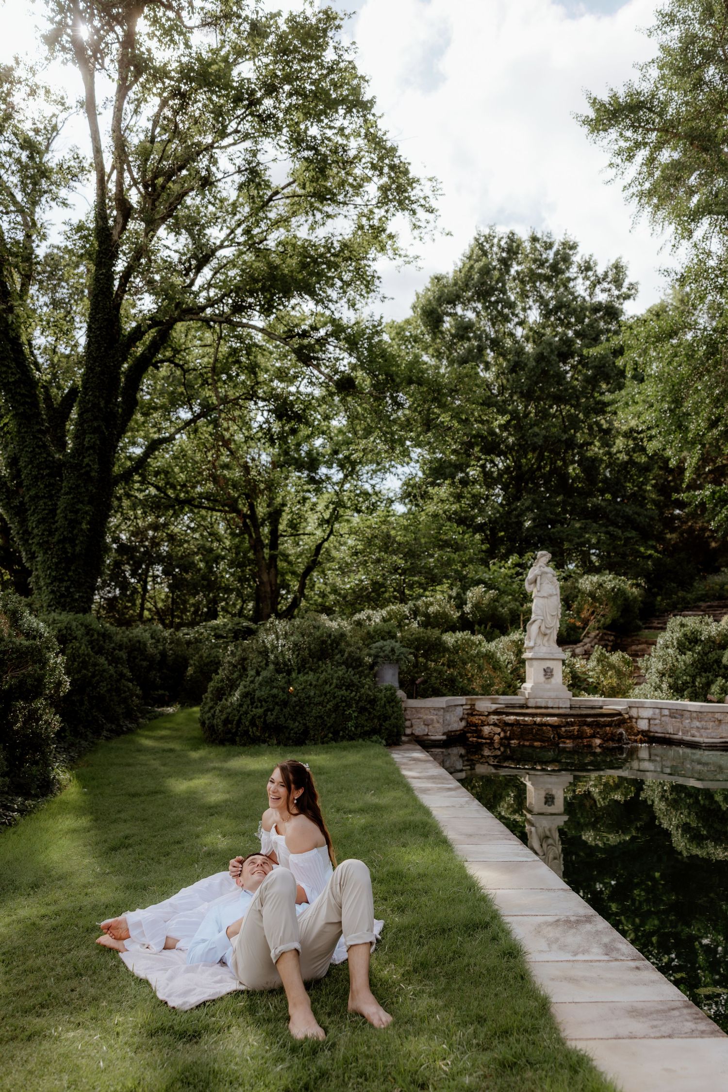 Two women relaxing on lush green grass beside a serene garden pond with a classical stone statue nearby.