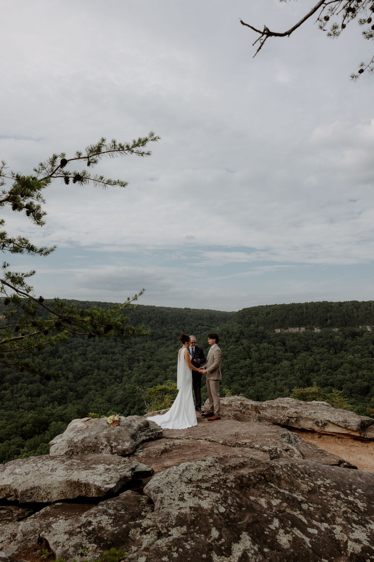 A couple exchanges vows on a rocky cliff overlooking lush green forested valleys during an outdoor mountain wedding ceremony.