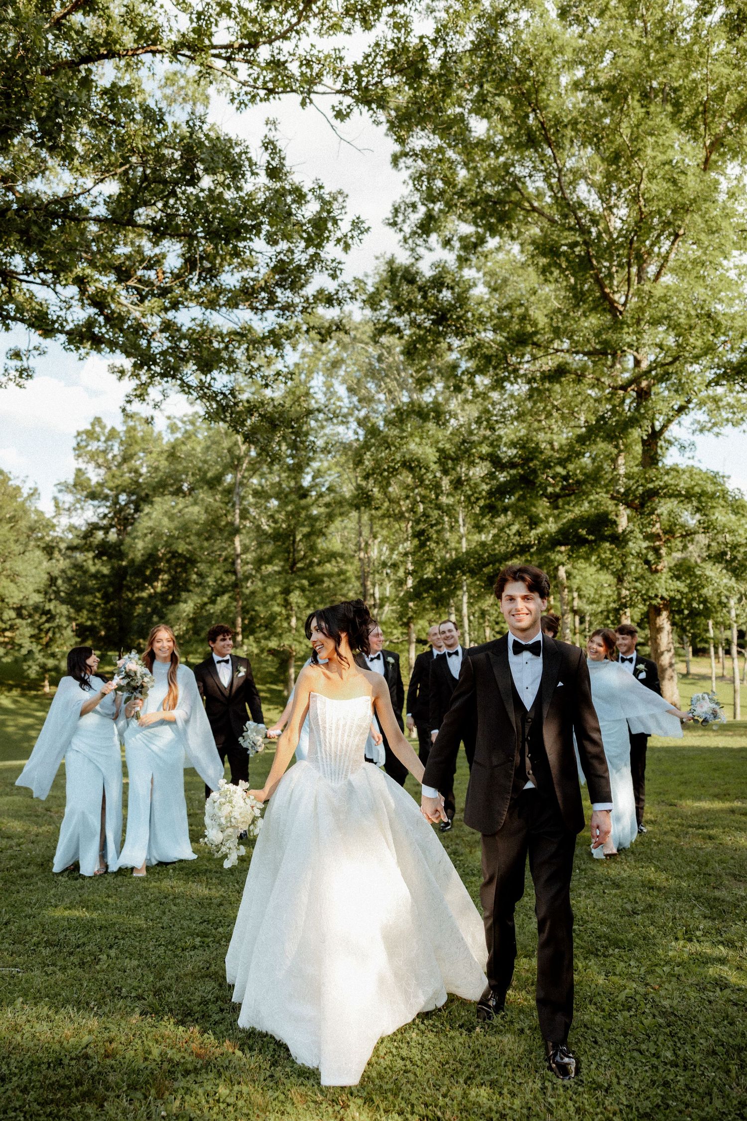 Bride and groom walk hand-in-hand through lush green park with wedding party following behind.