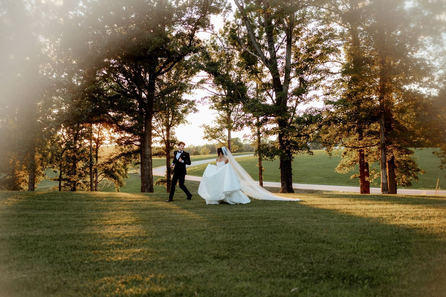 Bride and groom walk across lush green lawn at golden hour, surrounded by tall trees at outdoor wedding venue.
