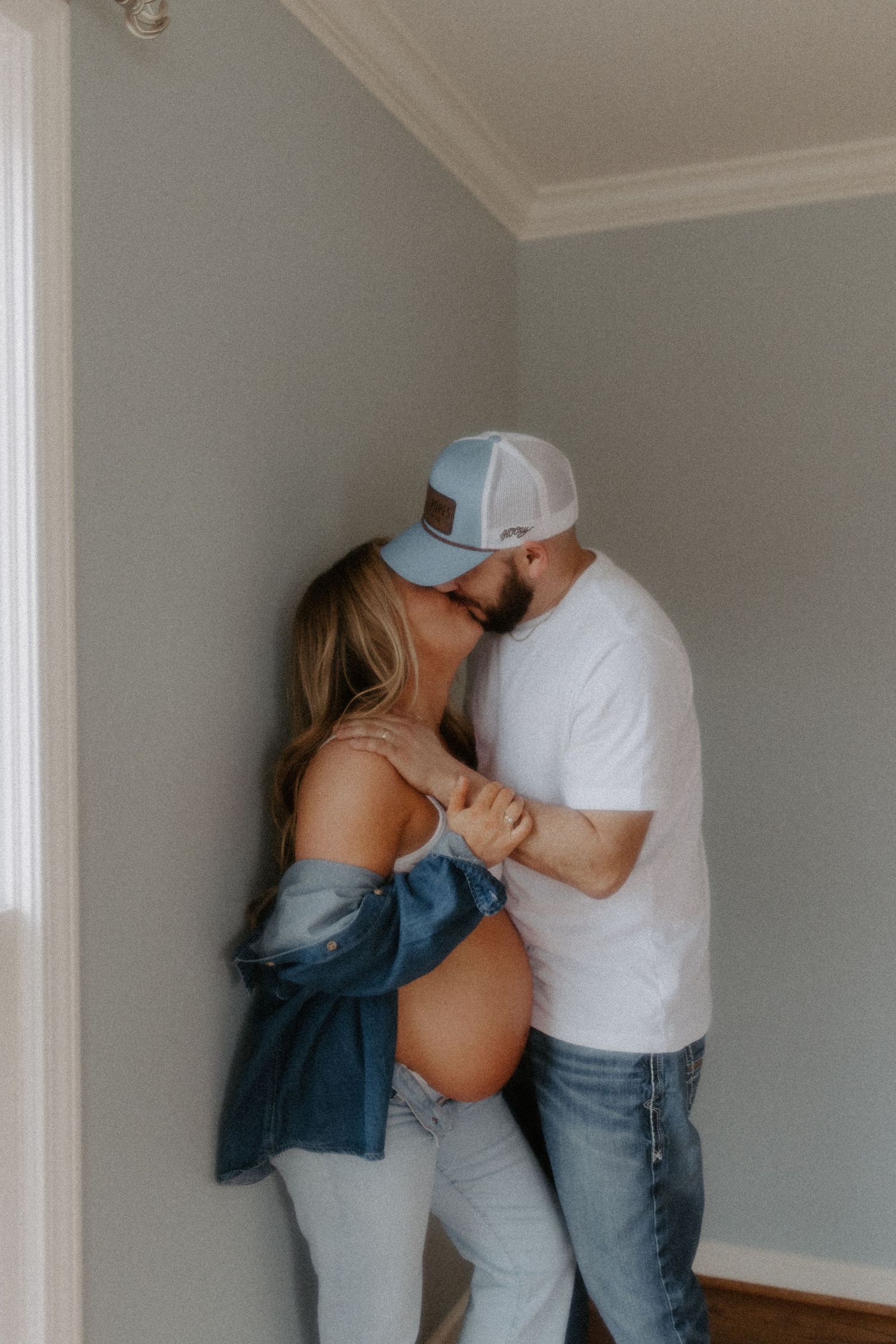 Pregnant couple sharing a kiss against a gray wall, woman in denim jacket with baby bump visible, man in white tee and cap.