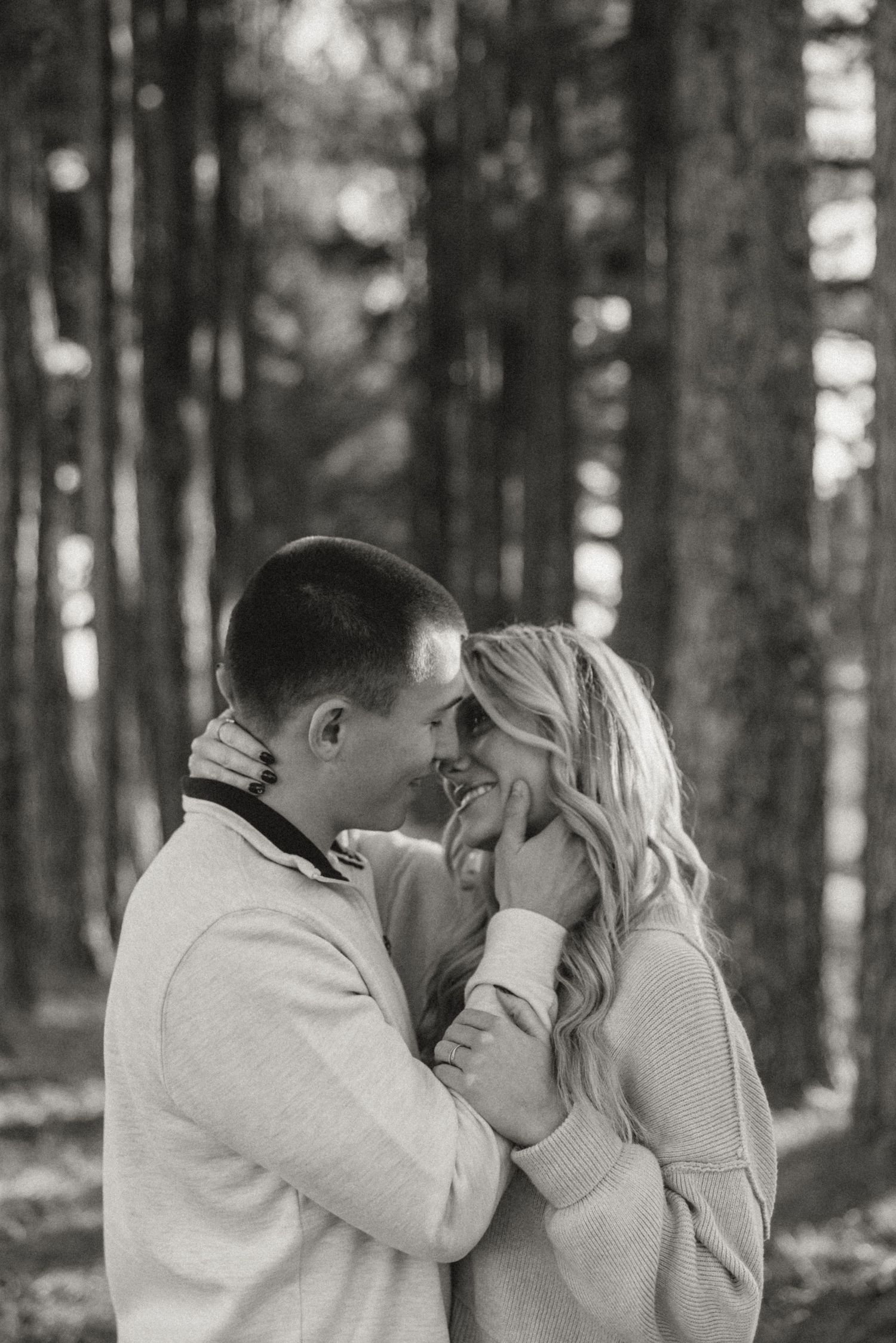 Black and white photo of a smiling couple embracing in a forest, touching foreheads romantically.
