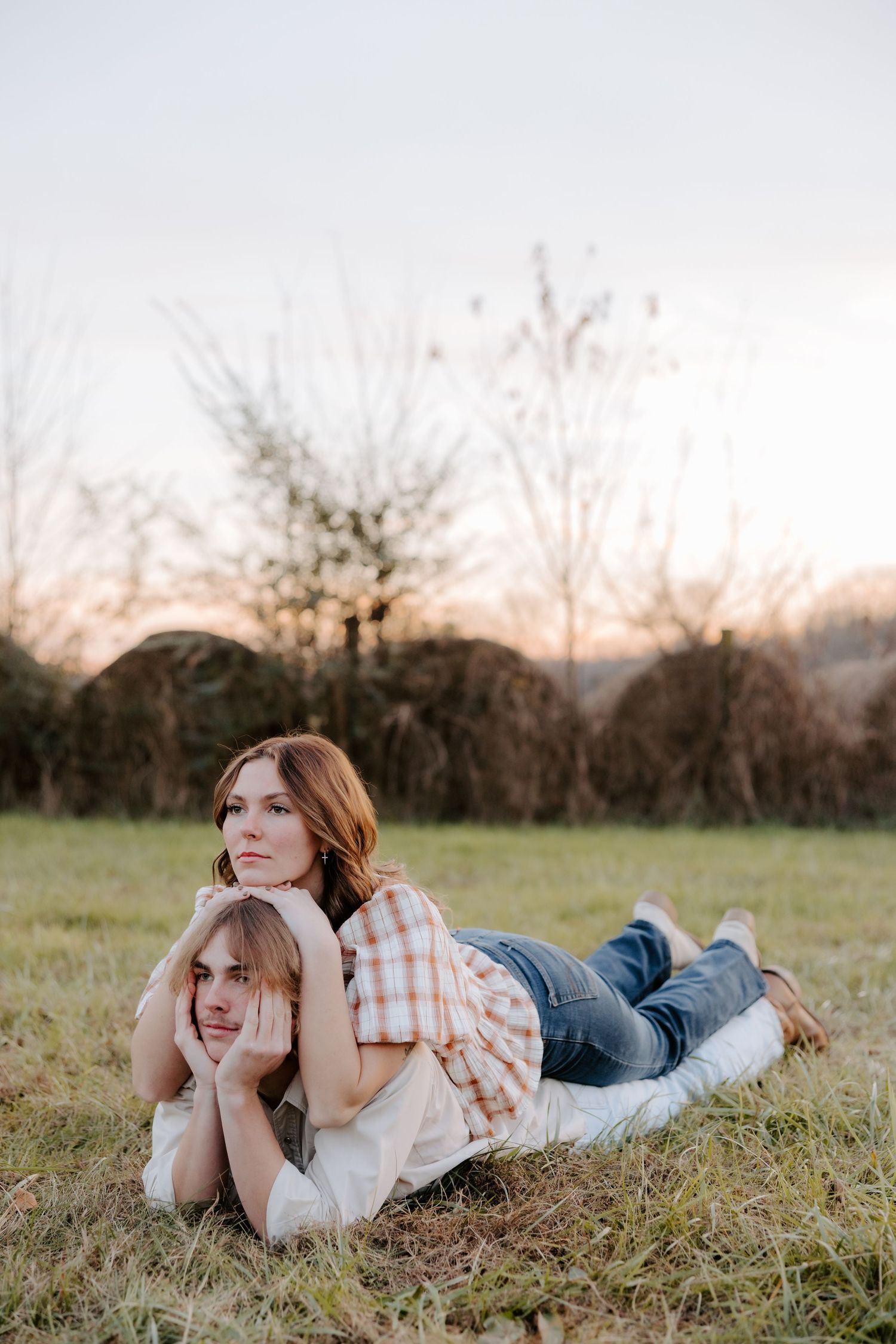 Couple lying on grass at sunset, woman resting on man's back, wearing jeans and plaid shirt, trees in background.