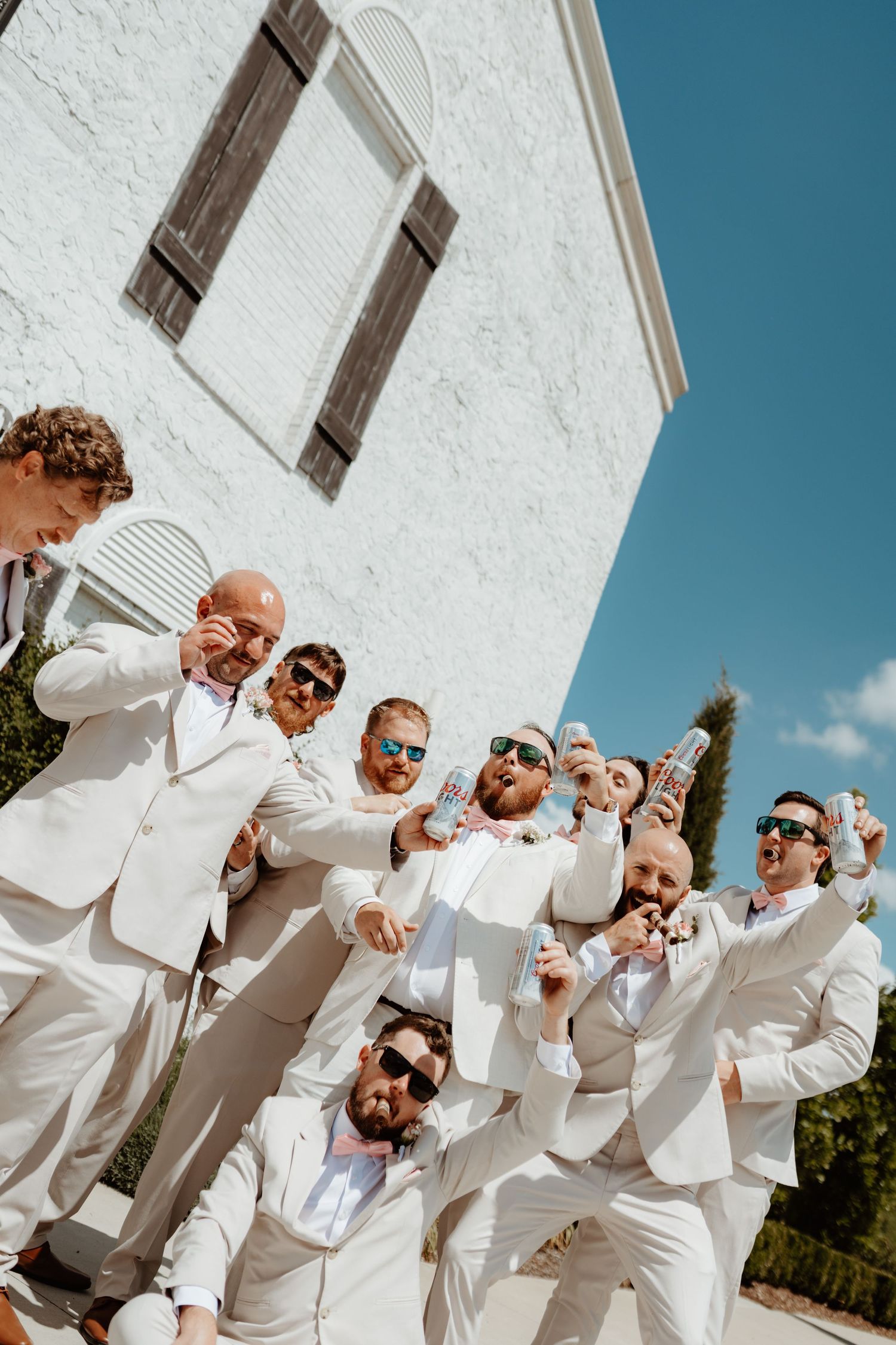 Groomsmen in white suits celebrate outdoors with drinks, wearing sunglasses near a white building under a clear blue sky.