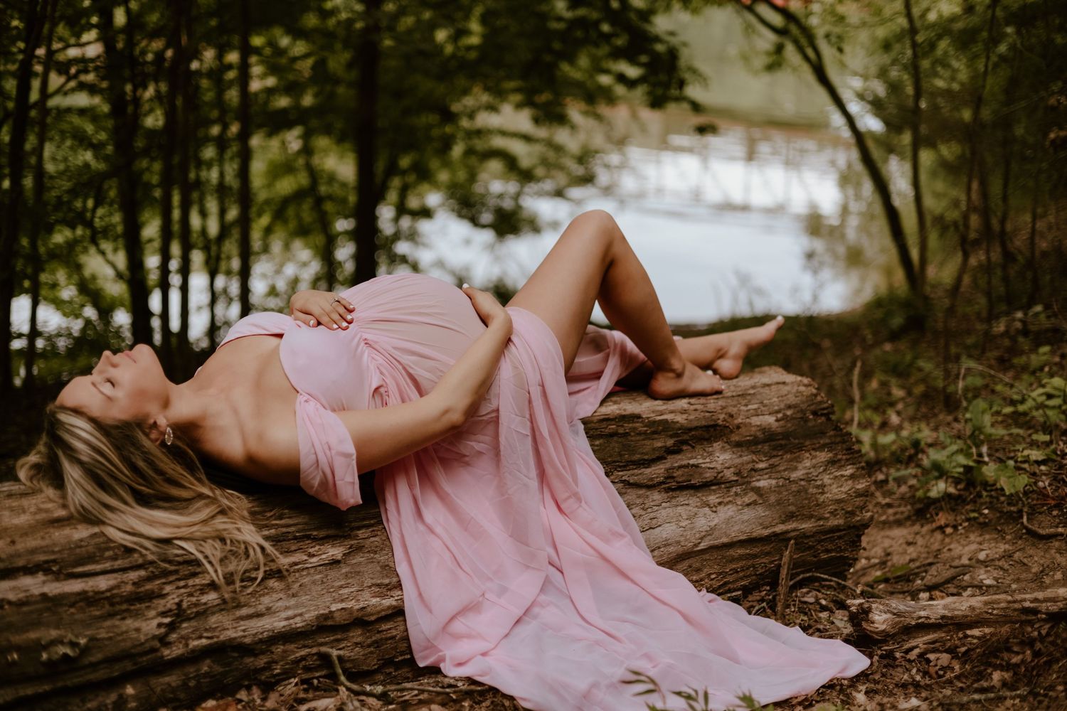 A person in a flowing pink dress lies gracefully on tree roots by a snowy forest lake.
