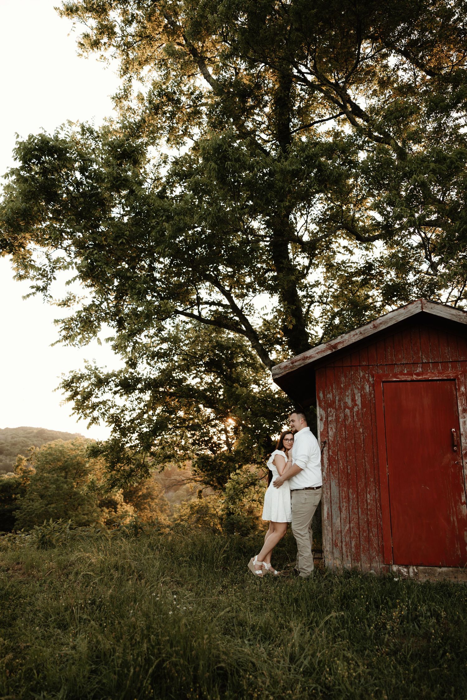 Couple embraces beside red barn under tall trees at golden hour.