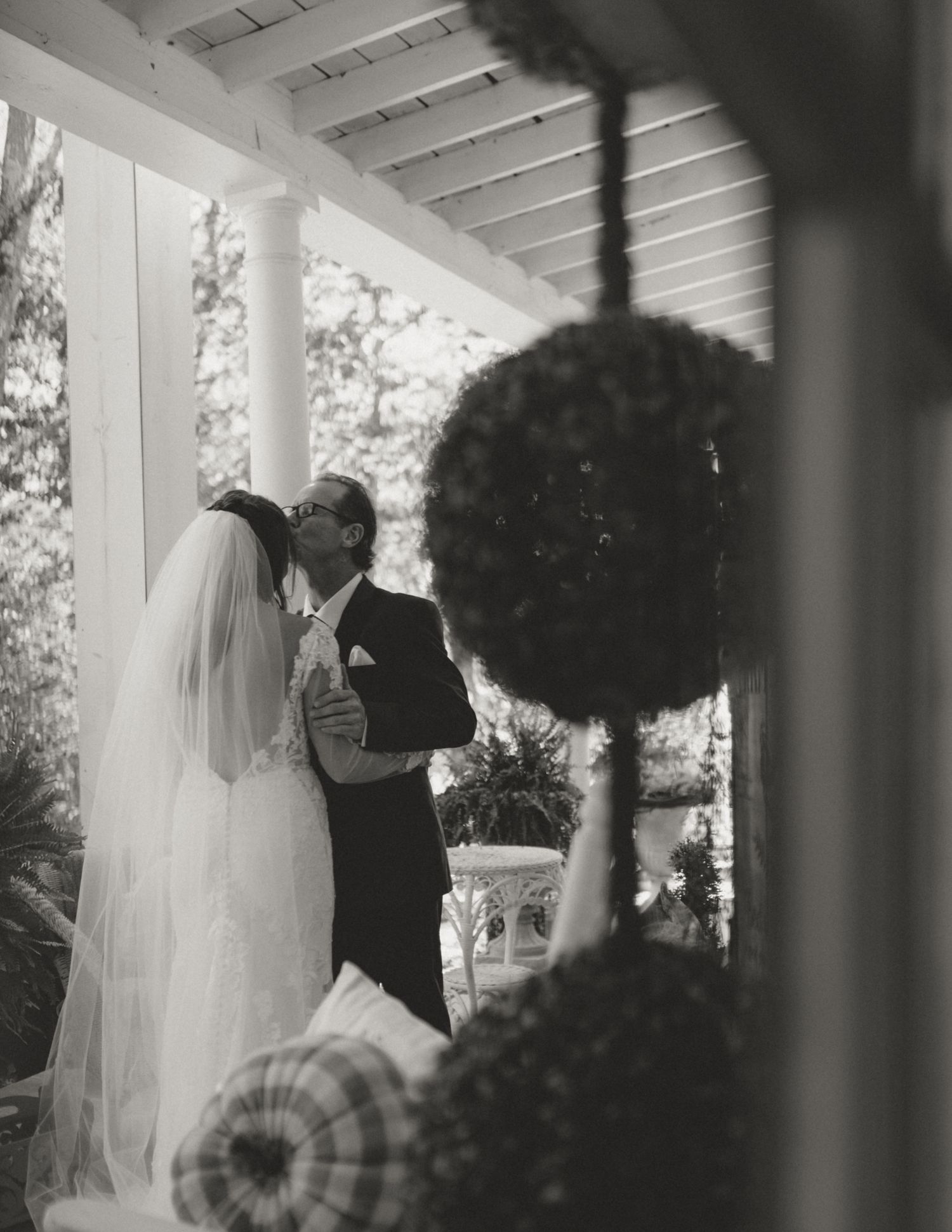 A black and white wedding ceremony photograph captured through decorative topiary on an elegant porch.