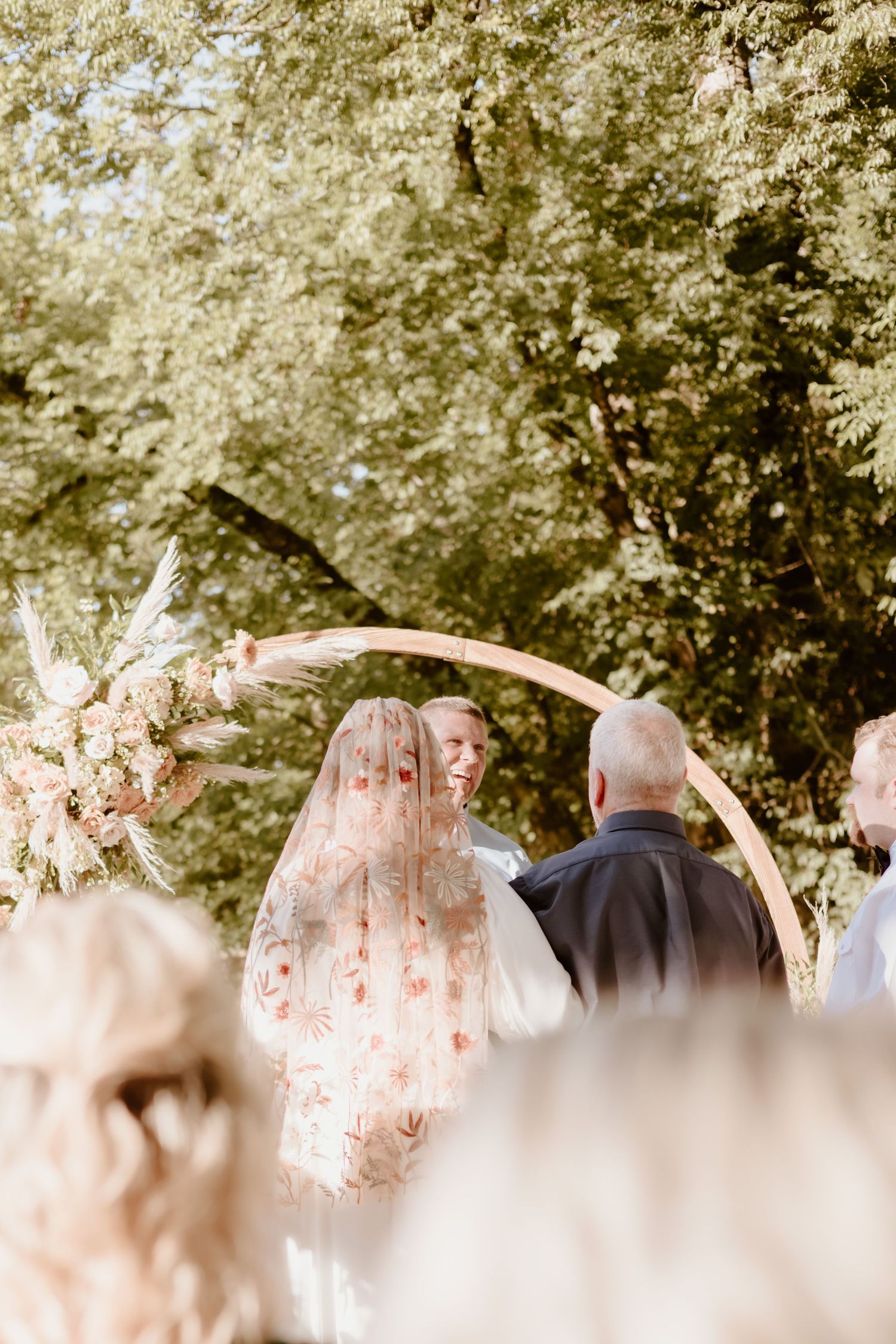 An outdoor wedding ceremony under a wooden arch adorned with dried flowers and pampas grass in a sunlit garden setting.