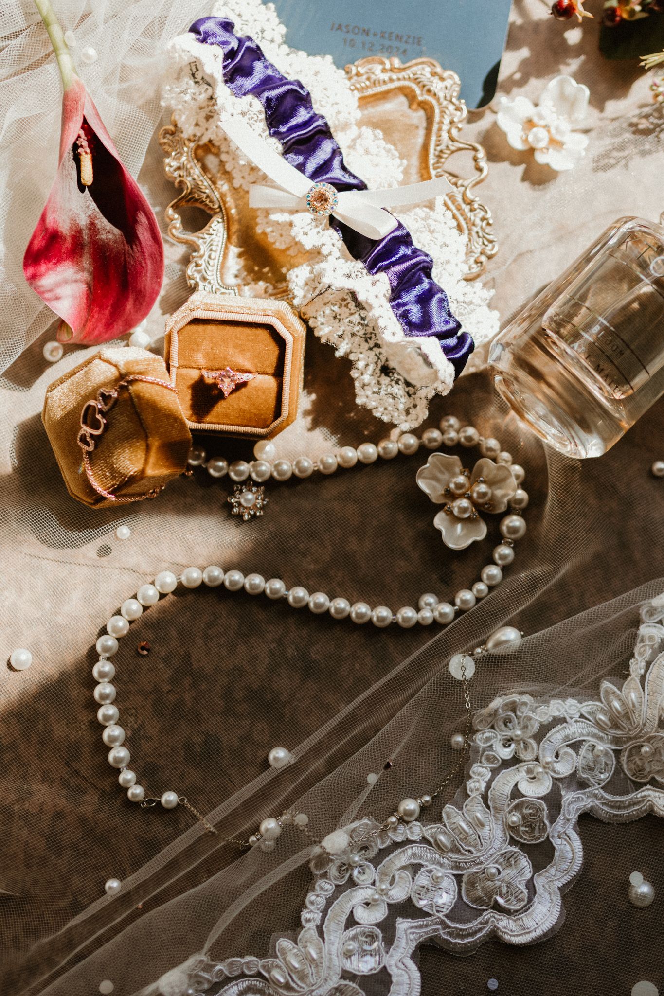 Wedding accessories including a pearl necklace, lace garter, ring box, perfume bottle and delicate lace trim arranged on dark surface.