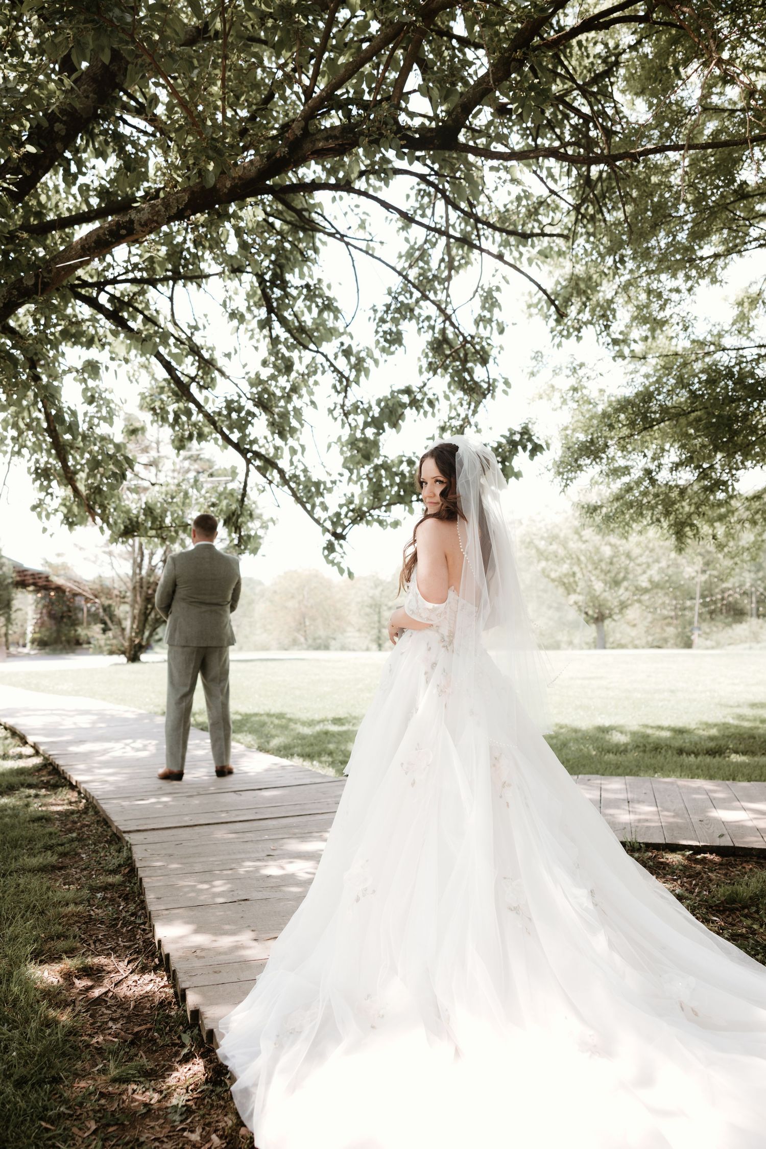 A flowing white wedding dress train trails elegantly along a tree-lined pathway in a romantic outdoor setting.