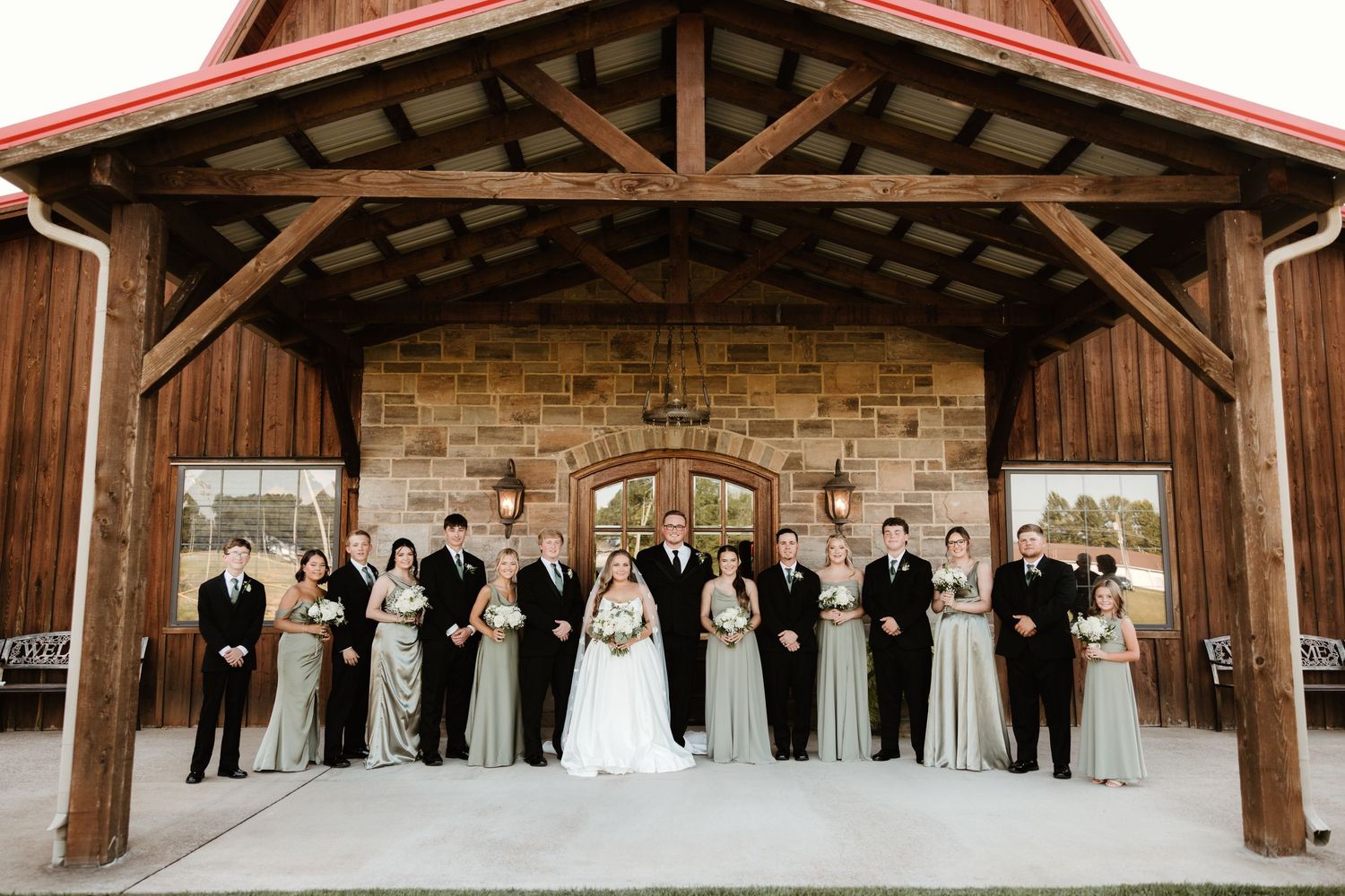 Wedding party poses together under a rustic wooden pavilion with stone accent wall.