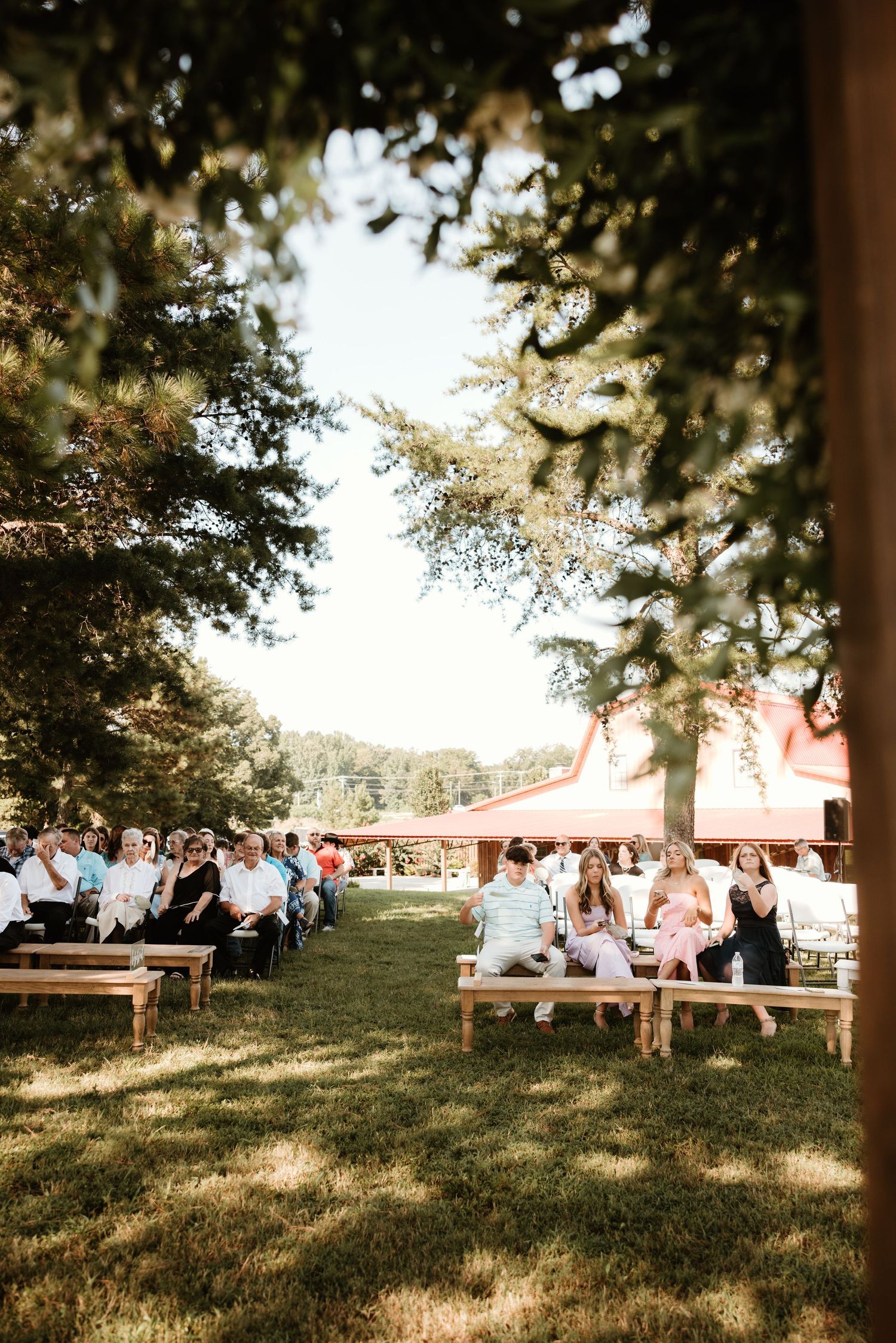 Rustic outdoor wedding ceremony seating with wooden benches under trees during golden hour in a garden setting.