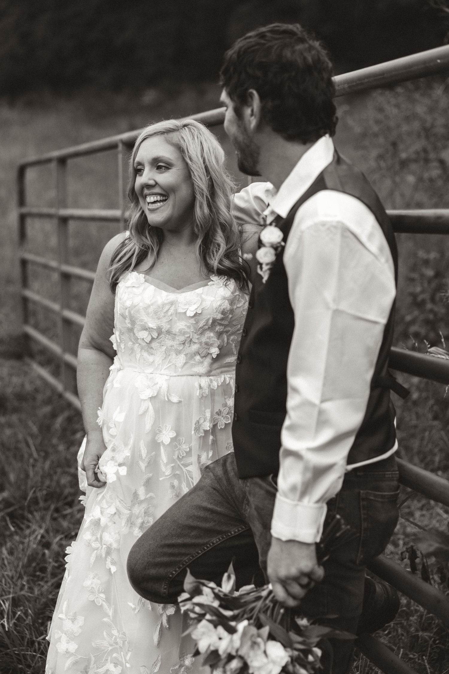 Black and white wedding photo of bride and groom sharing a joyful moment against a rustic fence in a rural setting.