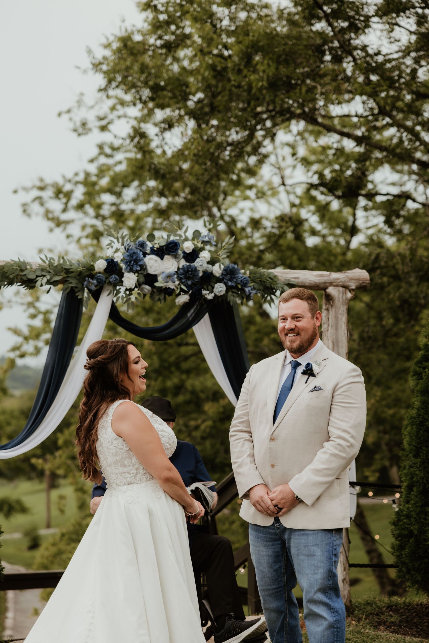 A couple exchanges vows under a floral arch decorated with white and navy draping at an outdoor wedding ceremony.