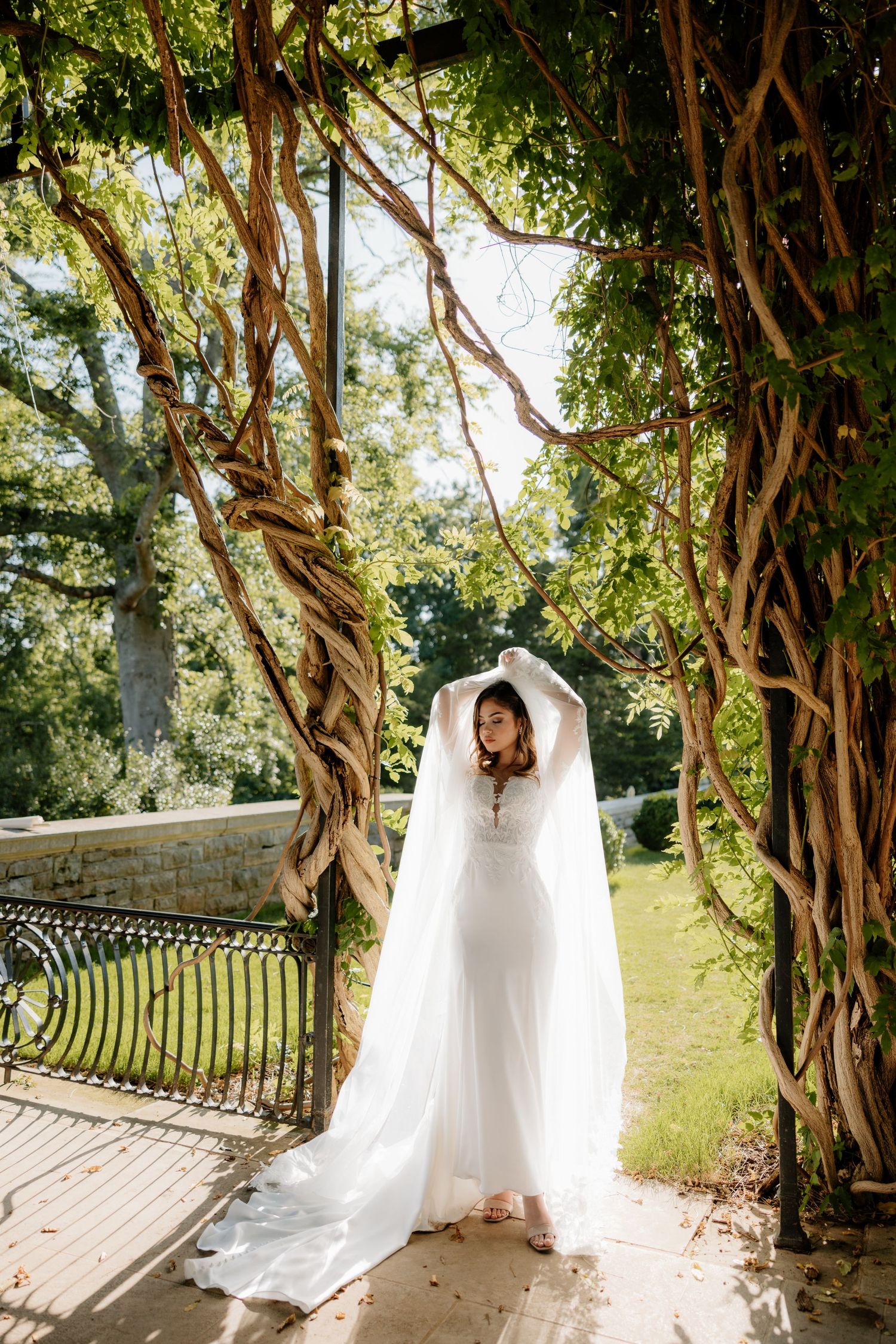 Bride in white gown and veil stands under vine-covered archway in lush garden, arms raised elegantly.