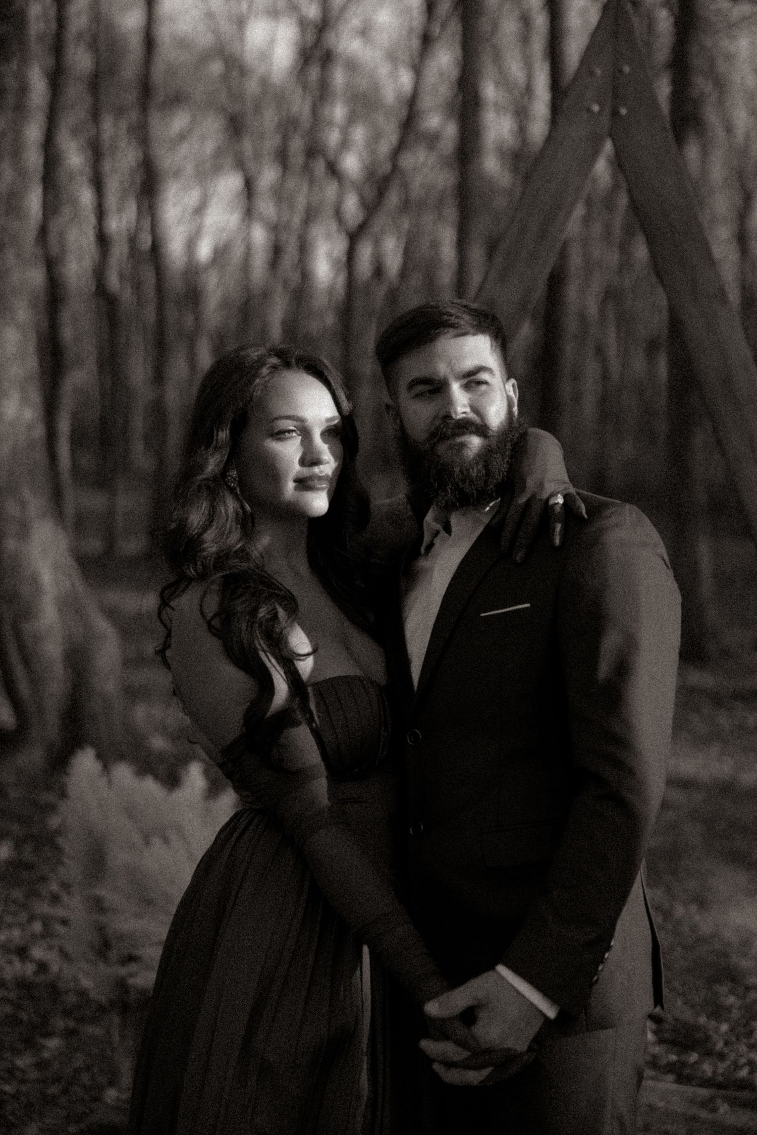 Black and white portrait of a bearded man in a suit and woman in an elegant gown embracing in a forest.