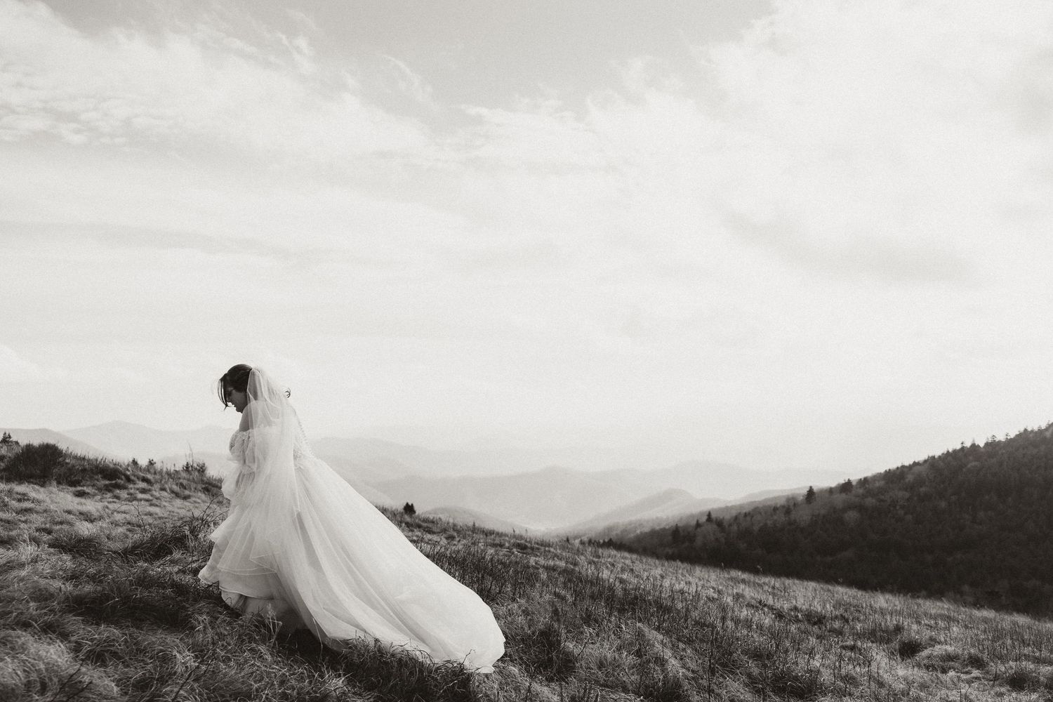 Black and white photo of a bride in a flowing gown and veil standing on a scenic mountain hilltop.