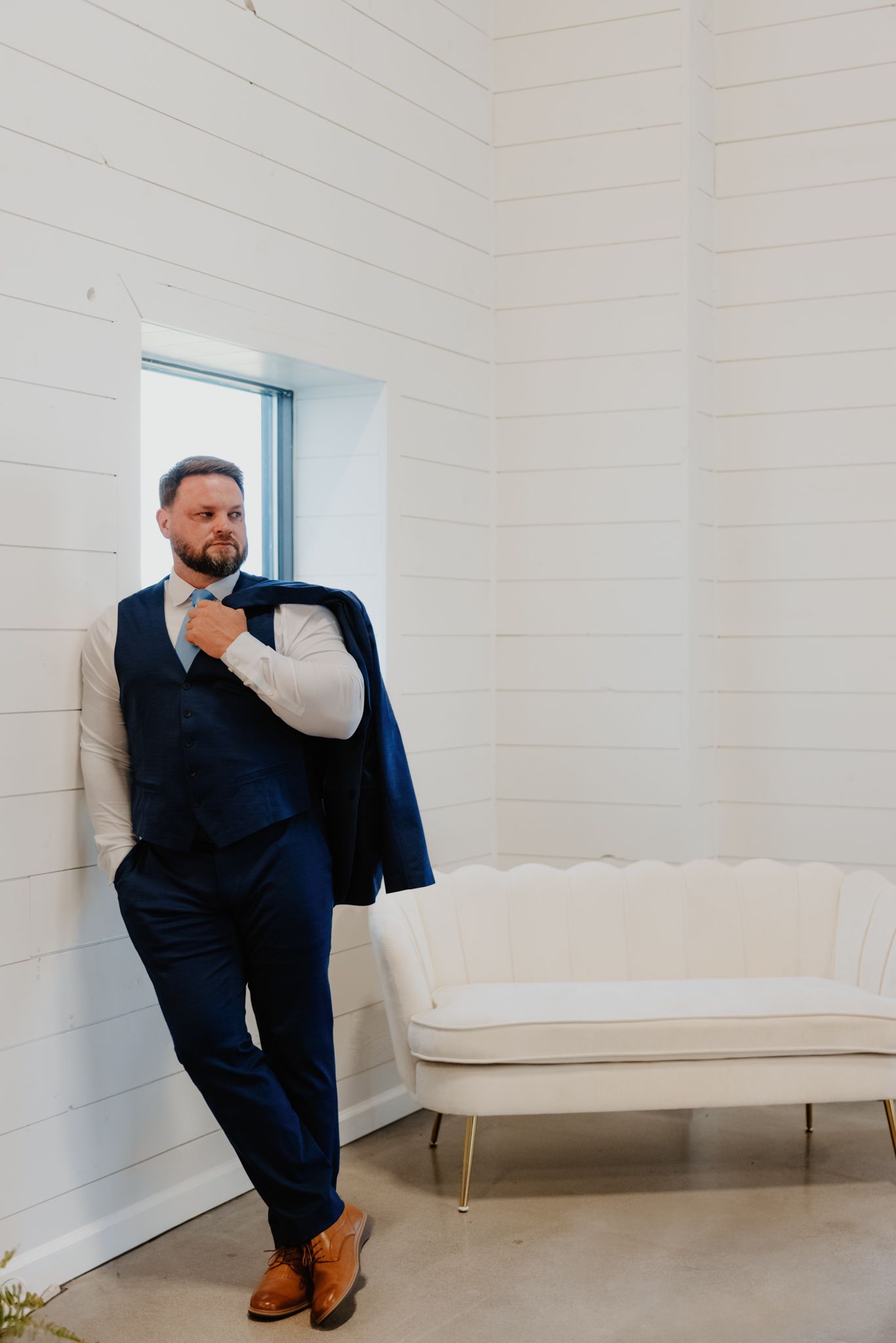 A person in a navy suit poses casually against a white wall beside a modern white settee near a window.