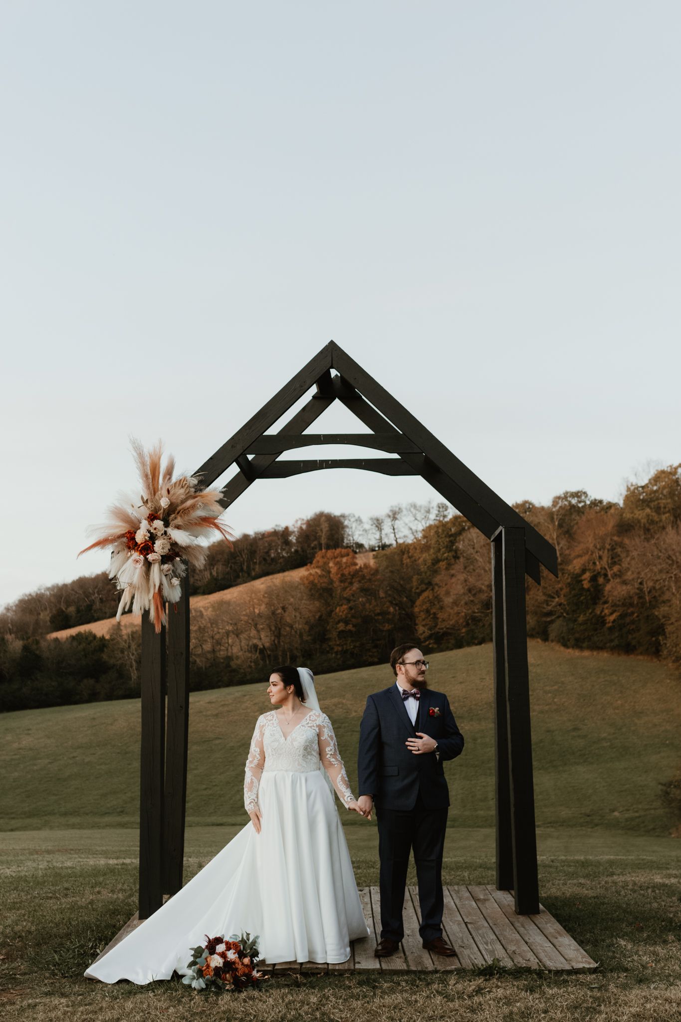 A romantic outdoor wedding couple stands beneath a rustic wooden archway against a scenic autumn hillside backdrop.
