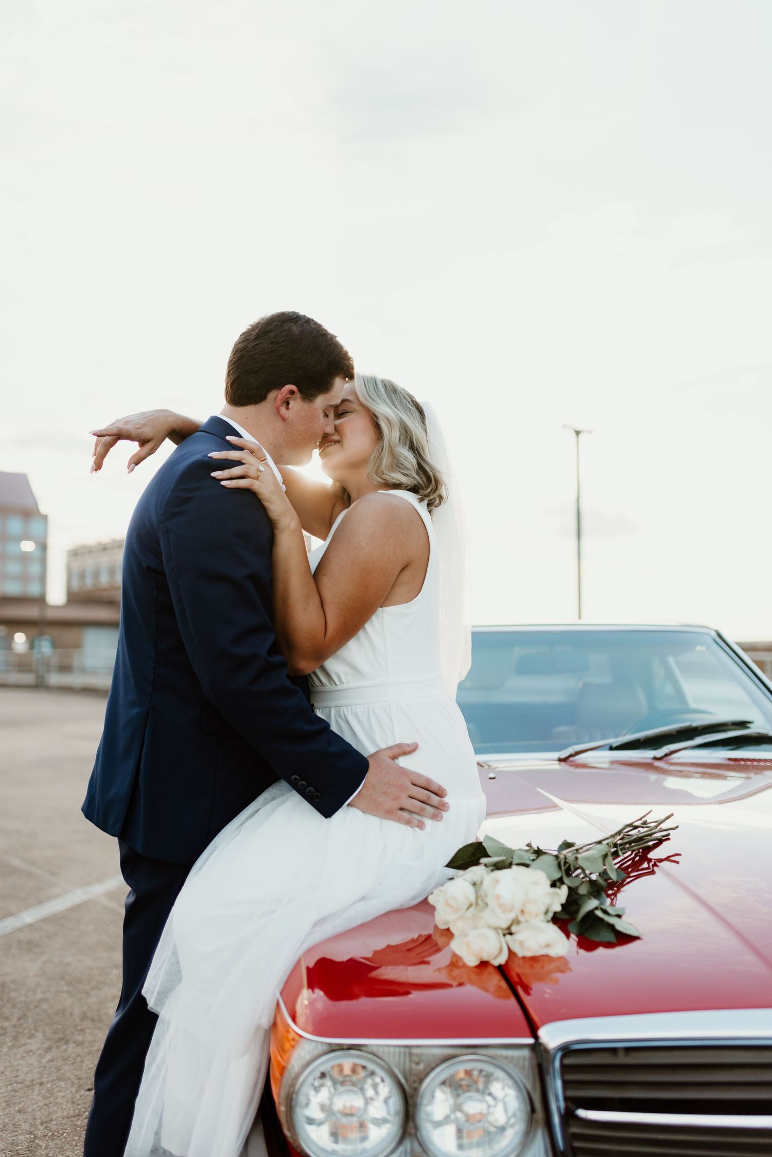 Romantic newlyweds share a kiss while leaning against a classic red car with white flowers on the hood.