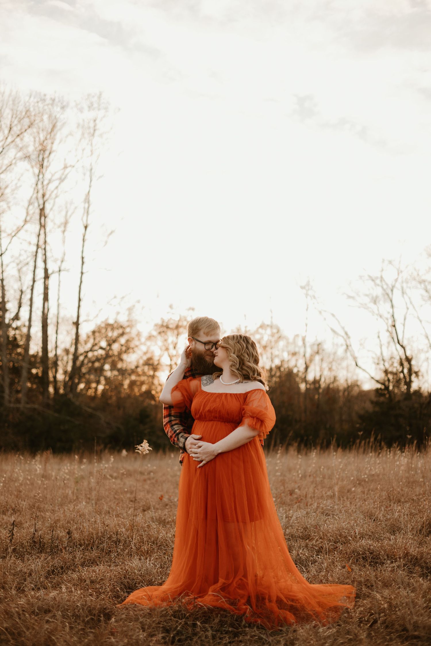 A couple embraces in a golden field at sunset, with the woman wearing a flowing rust-orange off-shoulder gown.