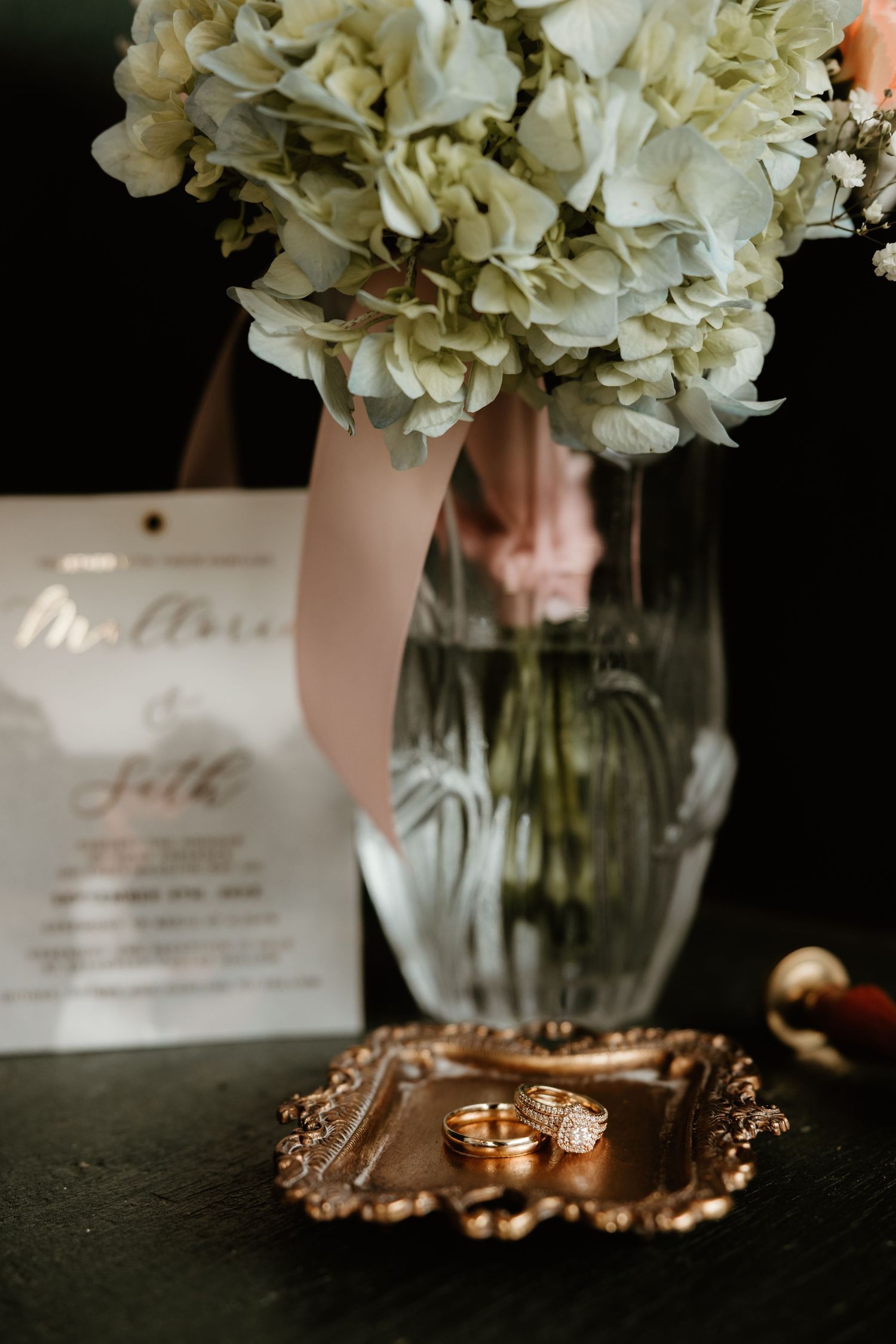 Wedding details showcase white hydrangea bouquets in glass vases with classic bronze accents and invitation cards.