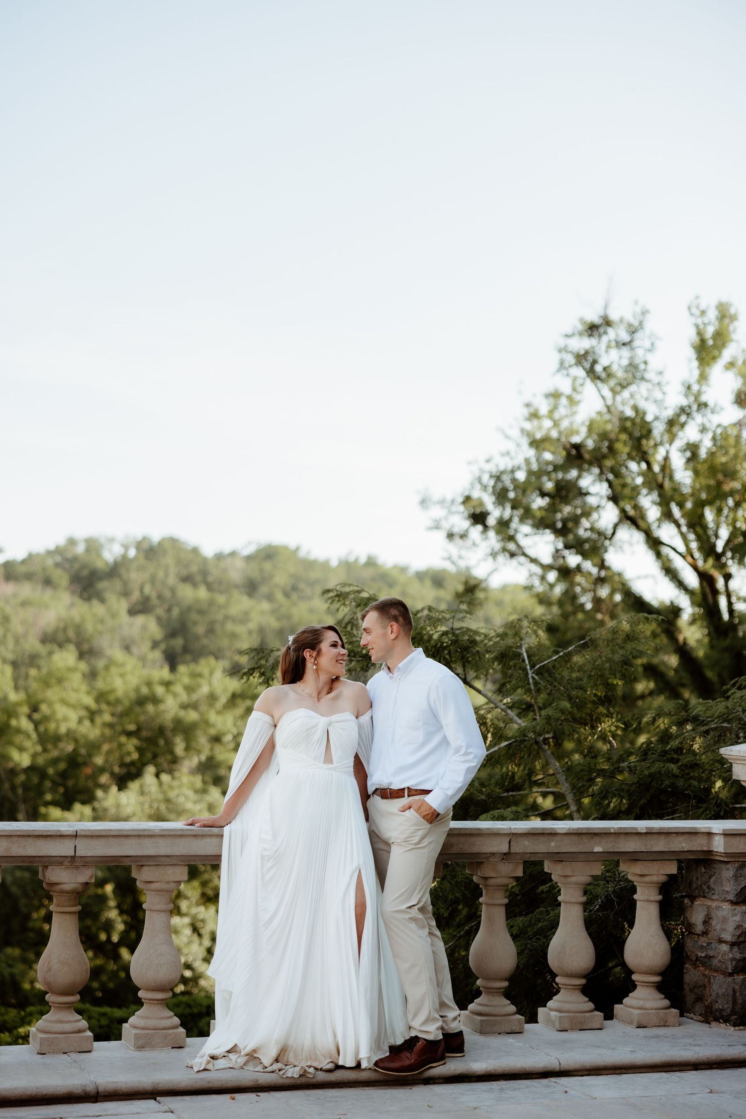 A bride and groom share a romantic moment on a stone balcony surrounded by lush green trees on their wedding day.
