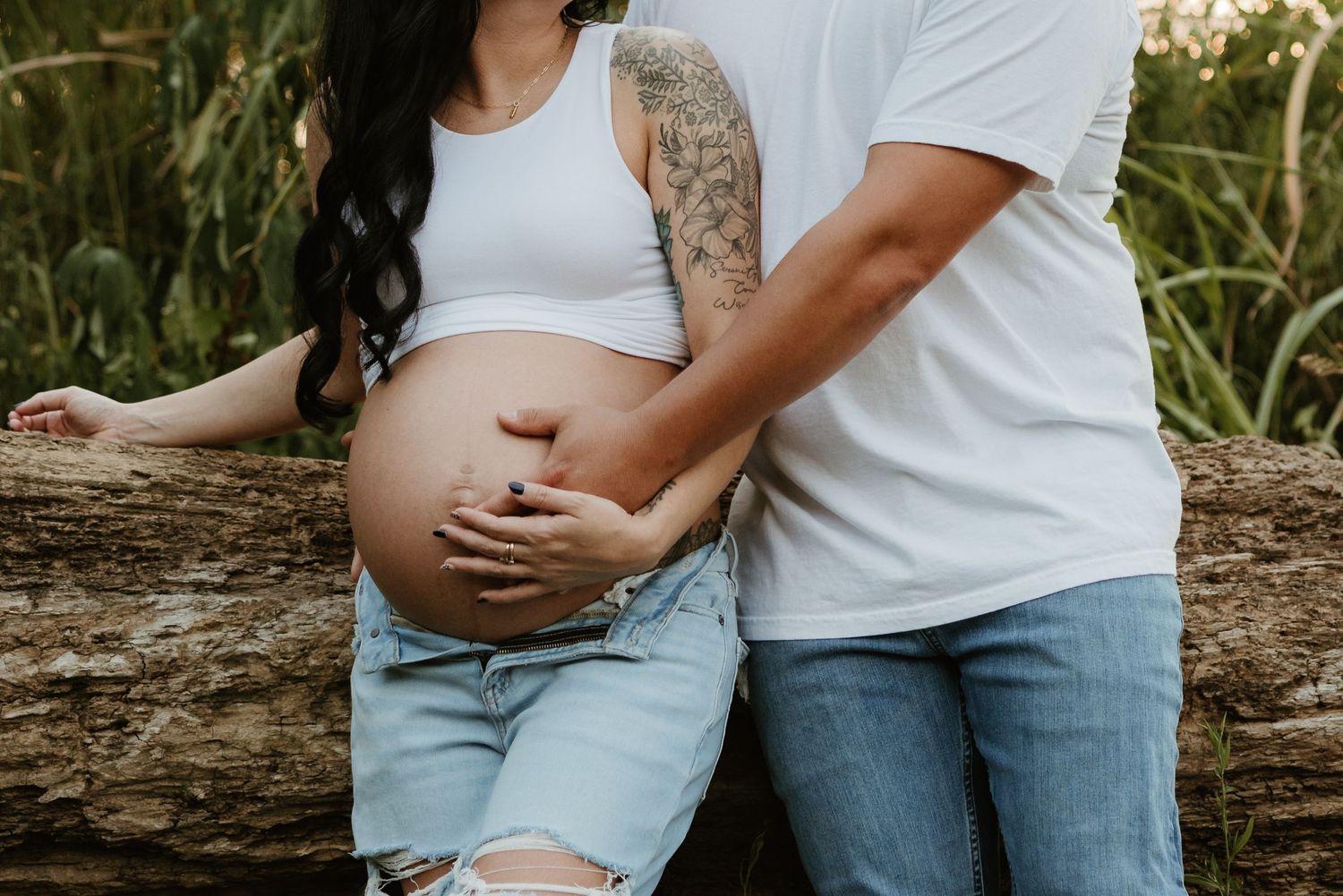 Two people in white shirts and denim rest on a log as hands cradle a pregnant belly in an outdoor maternity photo.