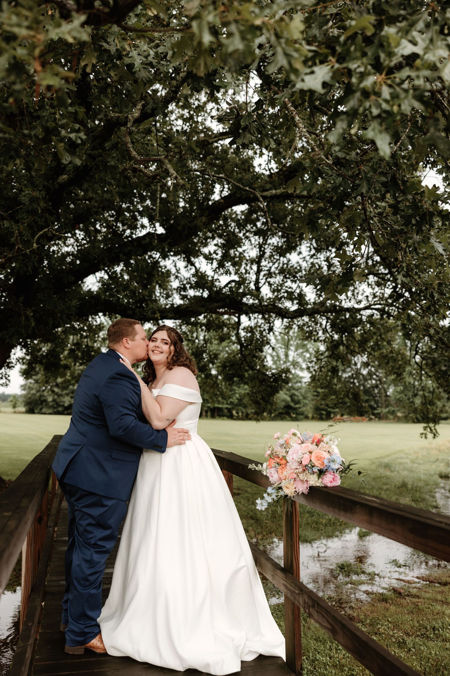 A romantic wedding couple embraces on a wooden bridge under oak trees with a pink bouquet resting nearby.