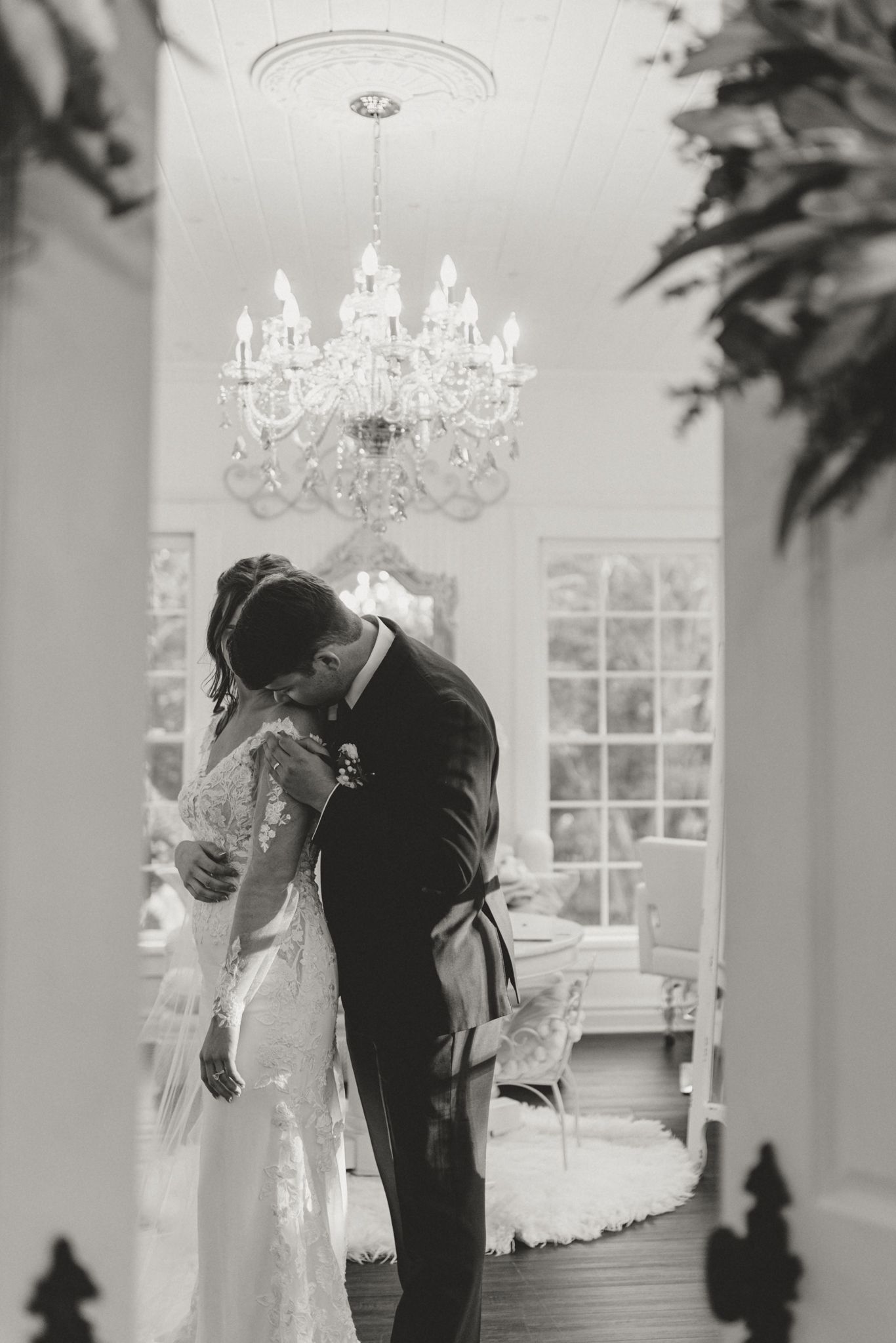 An intimate wedding moment beneath an elegant crystal chandelier in a bright white interior room.