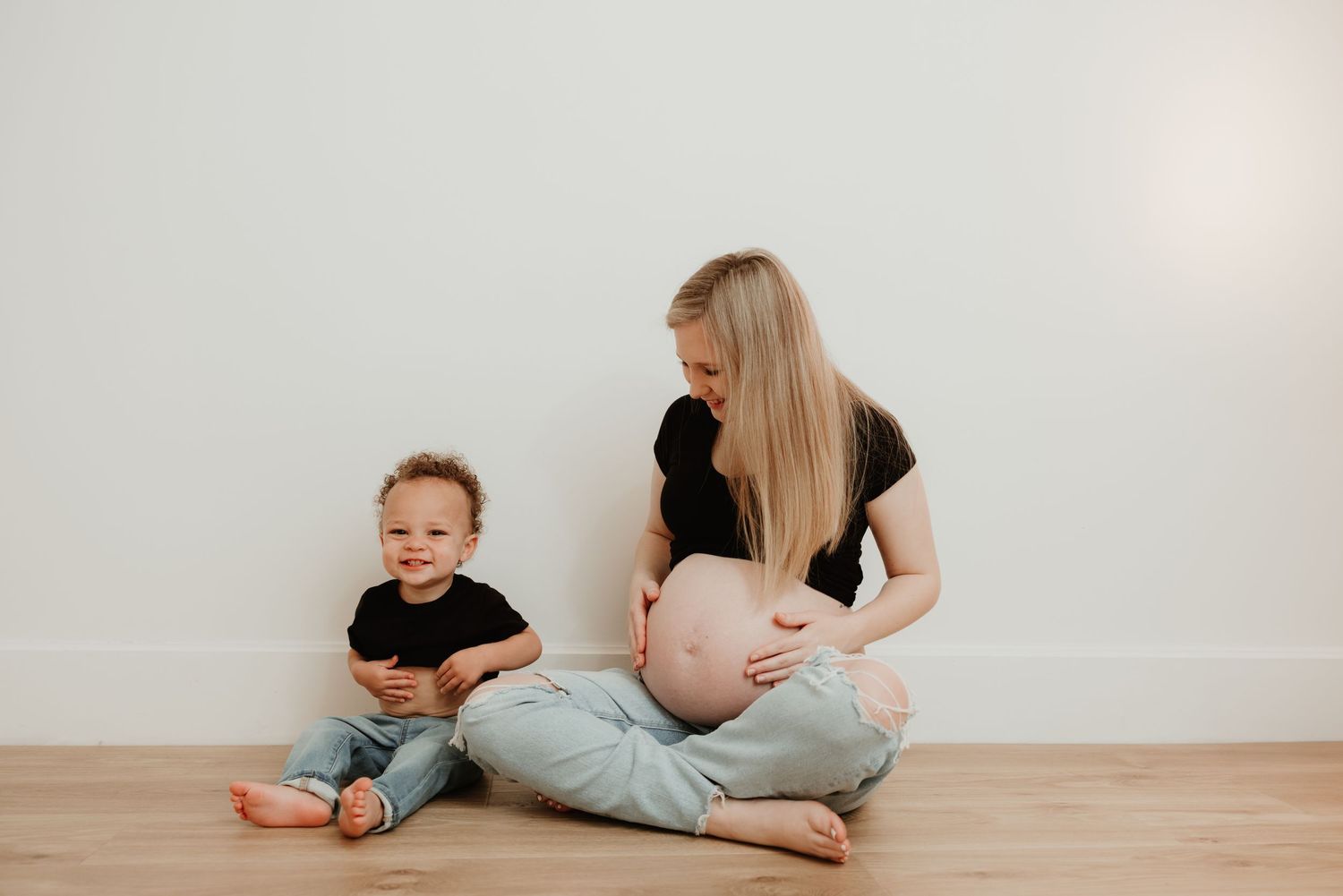 A pregnant woman and child sit together on a hardwood floor against a white wall, both wearing black tops and jeans.